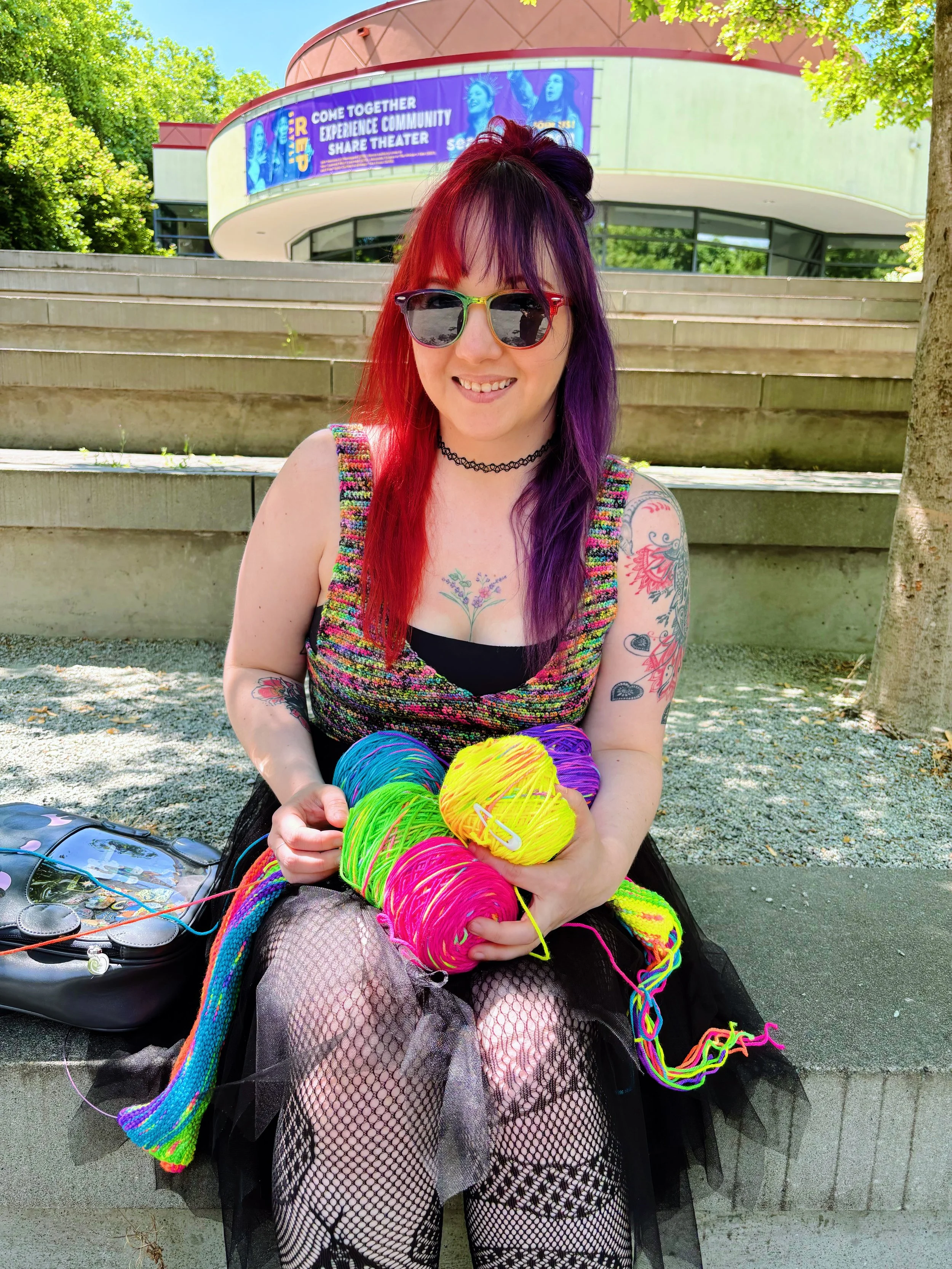 A woman with red and purple hair, wearing sunglasses and a black choker, smiling while sitting on steps outside. She is holding colorful yarn in her hands, wearing a rainbow-colored sleeveless top and fishnet tights. Behind her are trees, a building, and a banner that reads 'Come Together Experience Community Share Theater.'