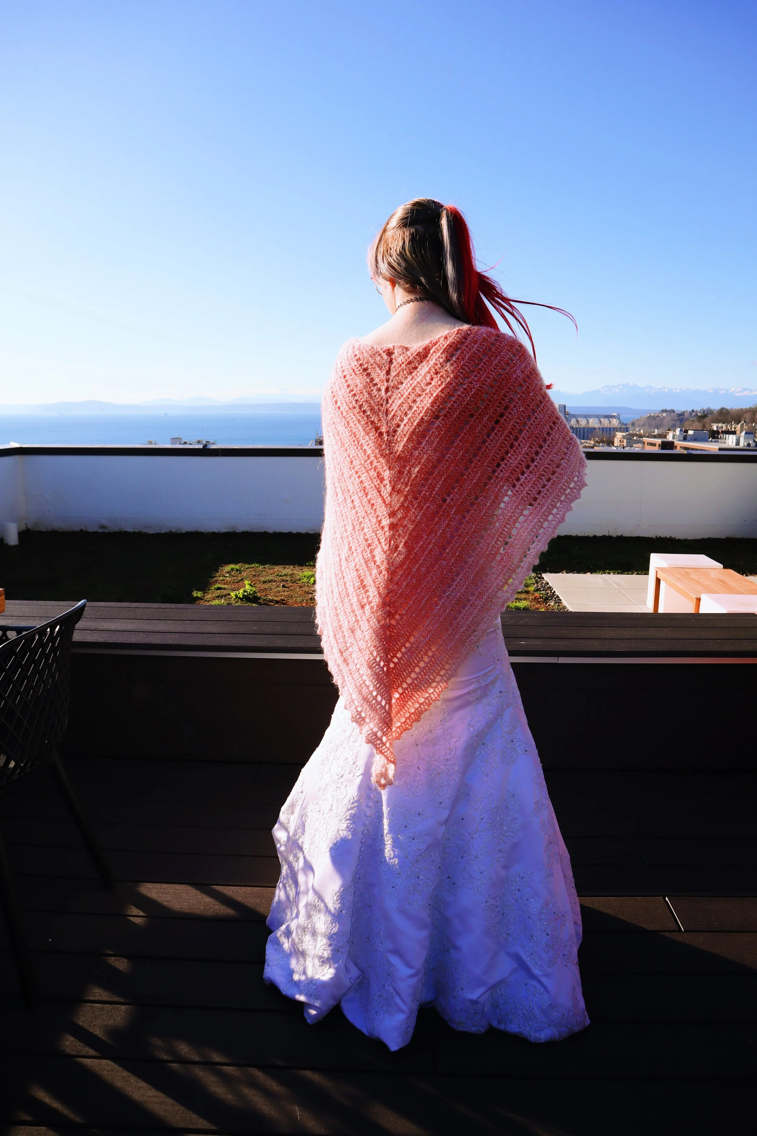 A person stands on a balcony, facing away, wearing a white lace gown and an orange and pink shawl, with a scenic view of mountains, water, and a clear blue sky in the background.