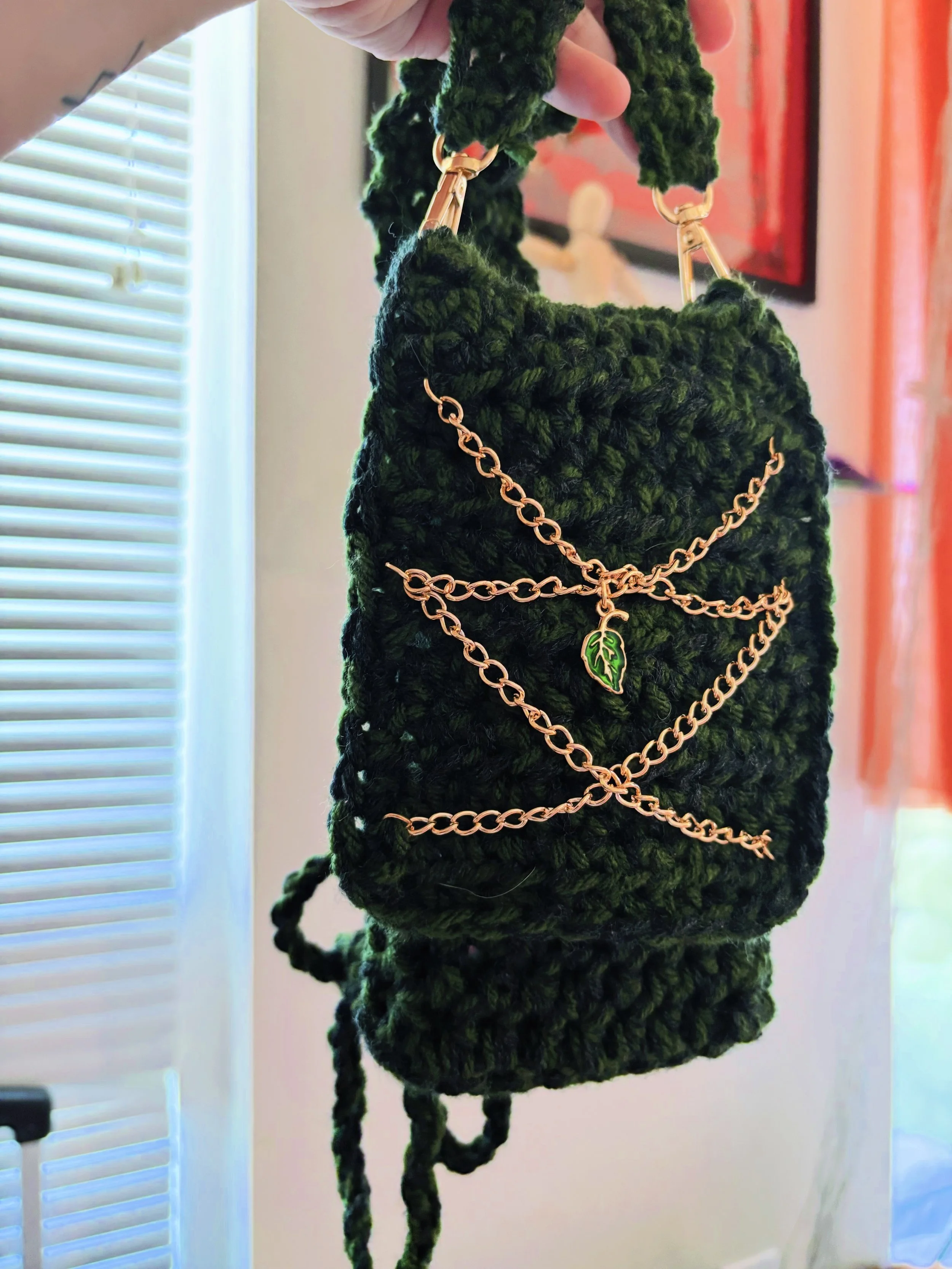 A person is holding a black crocheted or knitted bag with gold chain straps, one of which has a green leaf pendant attached. The background shows a window with horizontal blinds and a blurred framed picture or mirror.