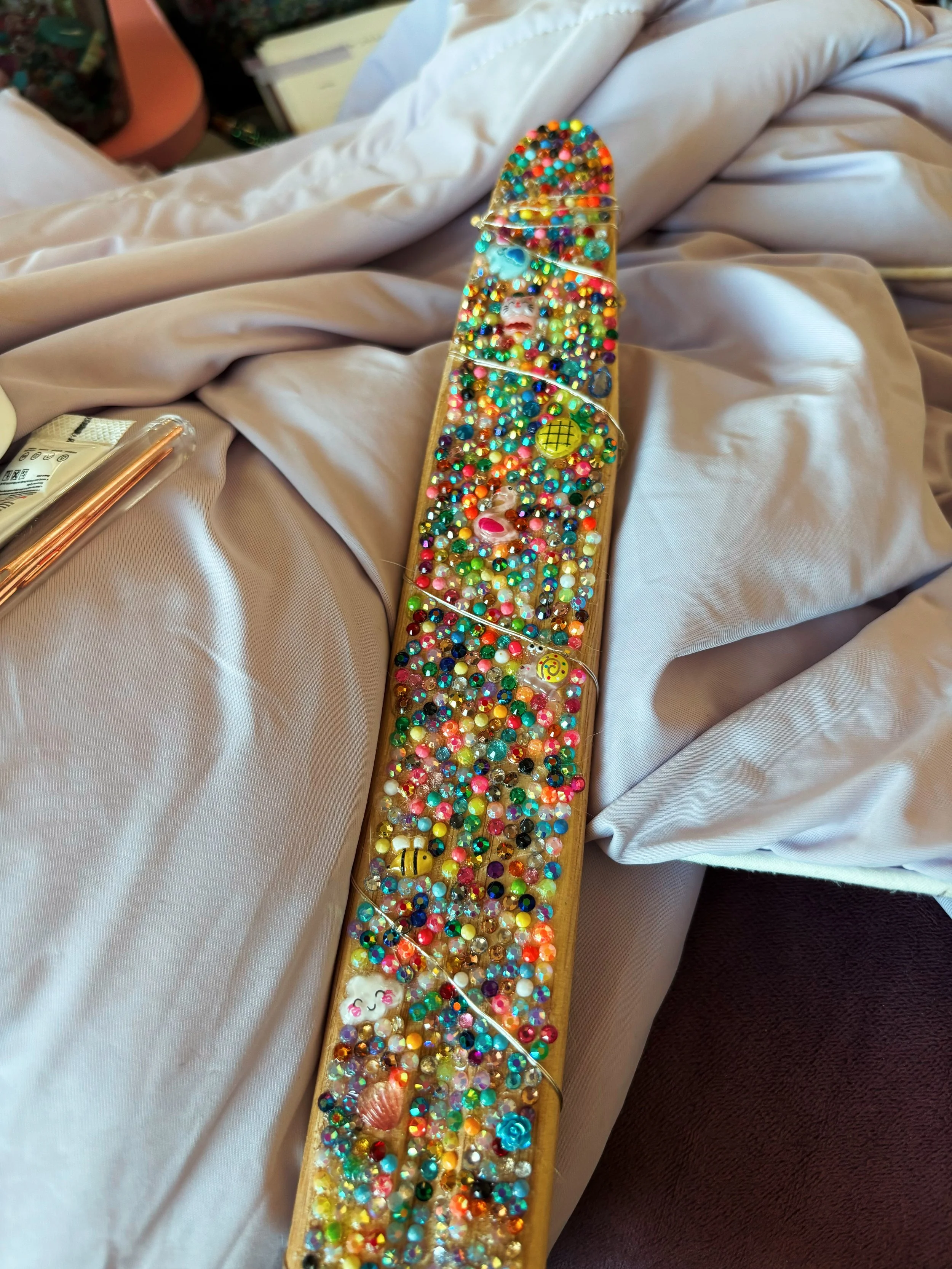 A felt paddle filled with colorful small beads, smiley face, and emoji decorations resting on satin fabric.