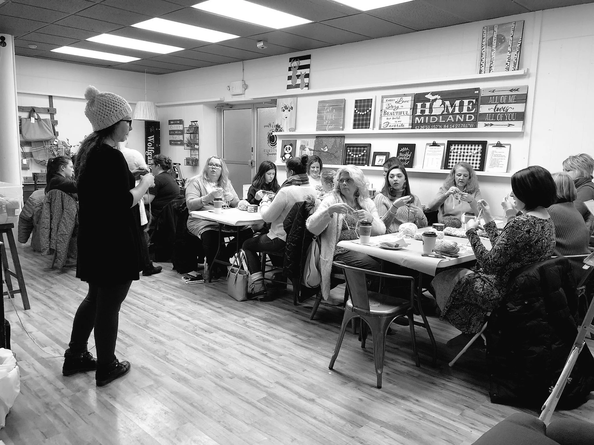 A group of gathered around tables in a cozy, decorated room, engaging in knitting and conversation. One woman stands wearing a knit hat, while others seated are holding knitting projects and drinks. The room features wall art with quotes and decorative plaques.