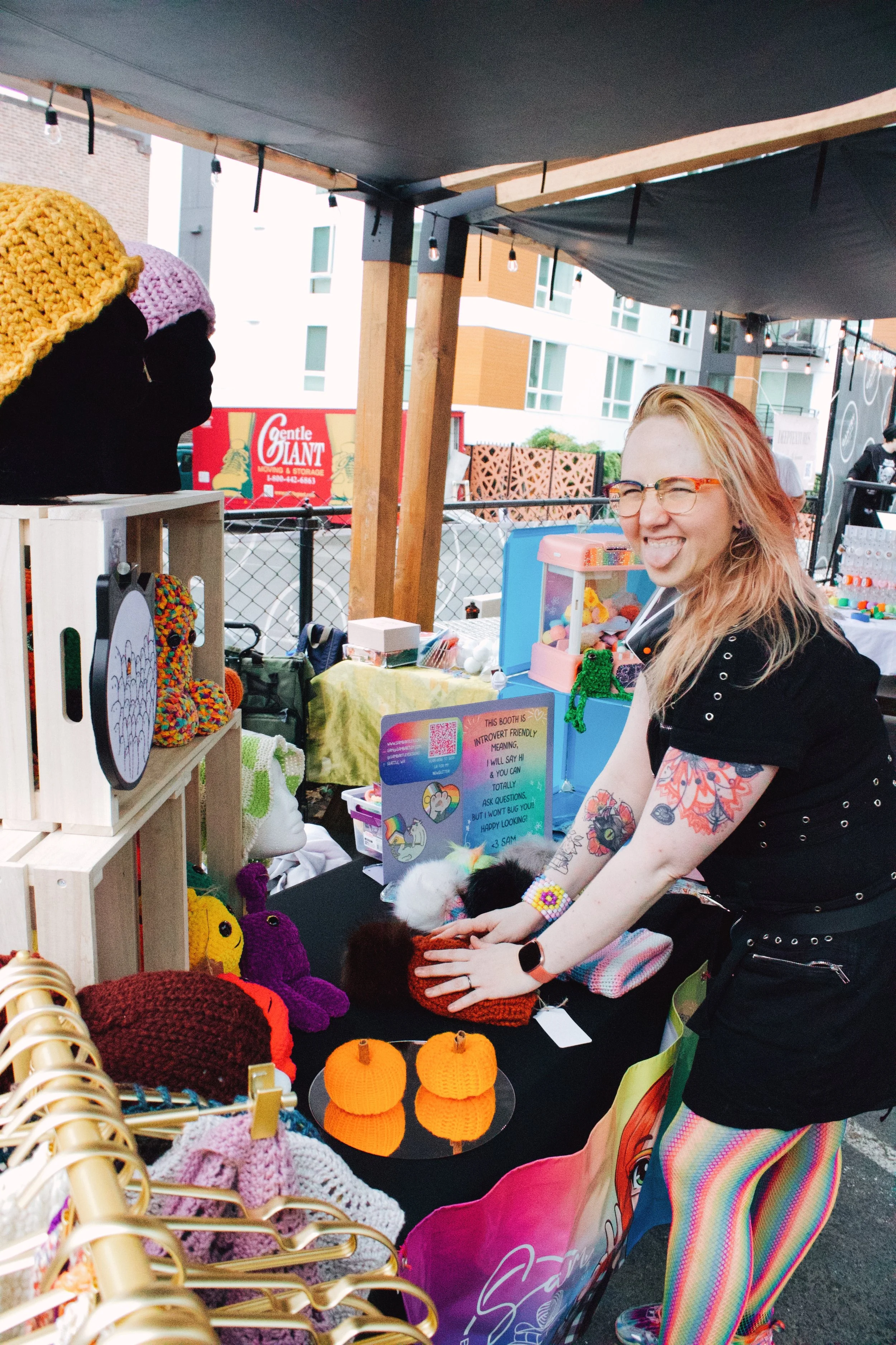 A woman with glasses and tattoos, wearing a black shirt with studded sleeves, is smiling and playing with yarn and stuffed animals at a craft booth at an outdoor market. The booth displays handmade knitted items such as hats and pumpkins, with a sign indicating it is a friendly, introvert-avoiding space.