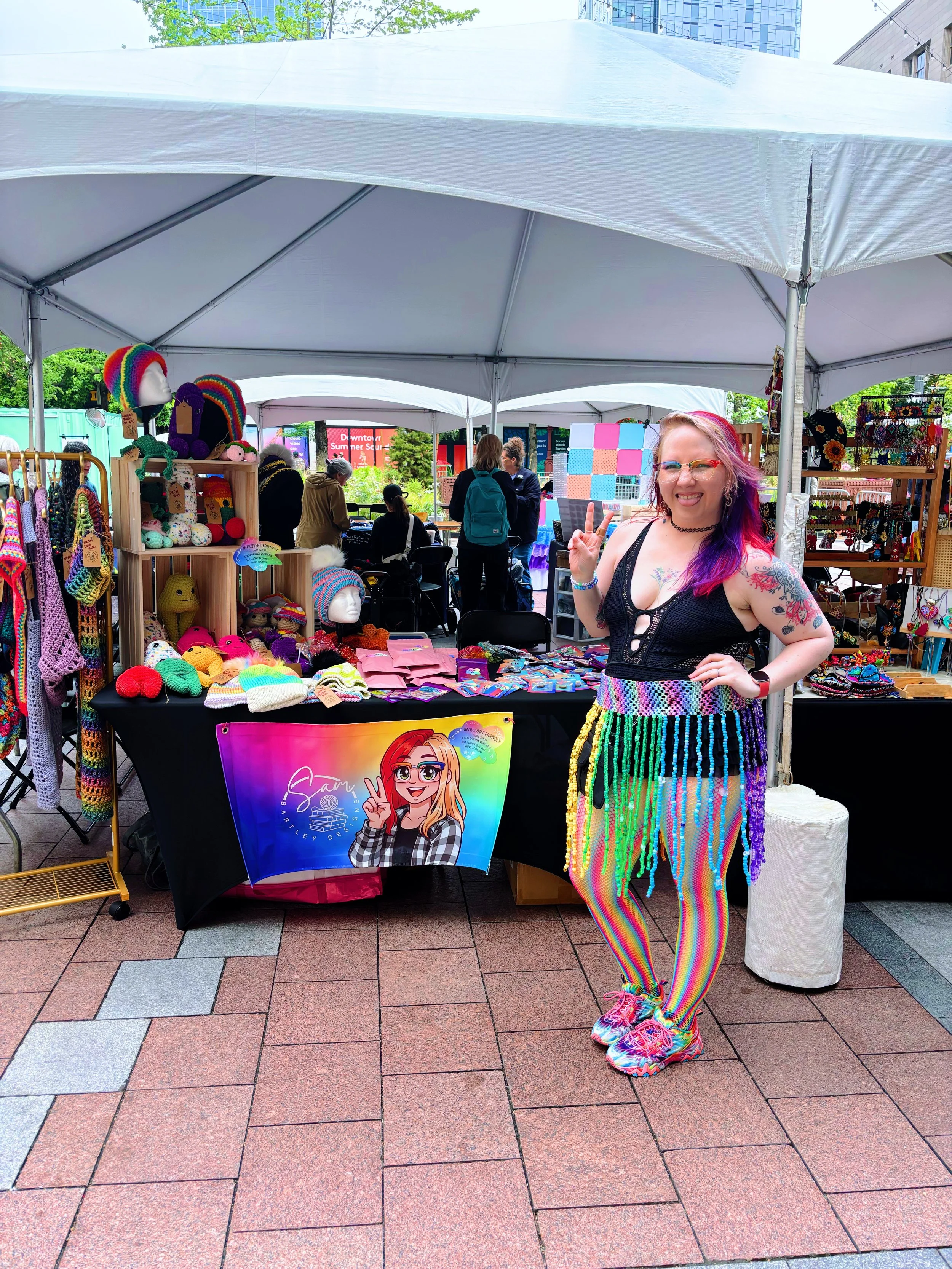 A woman with colorful hair and rainbow rainbow striped pants making a peace sign at a vendor booth selling knitted accessories and jewelry at an outdoor market.