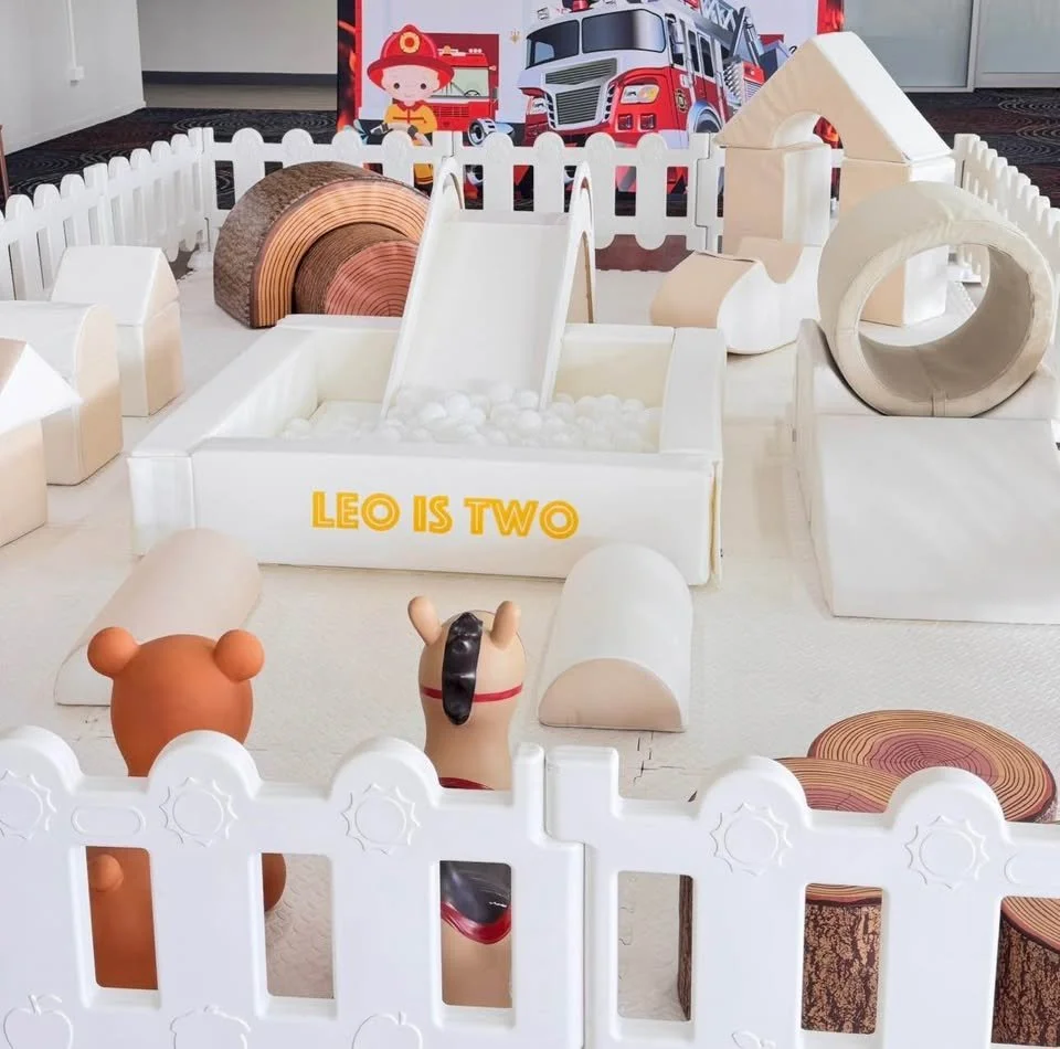 A small indoor play area with white plastic fencing, featuring a slide, a ball pit labeled 'Leo is Two', and various wooden and foam structures. There are toy animal figures, including a bear and a horse, and a colorful cartoon fire truck backdrop.