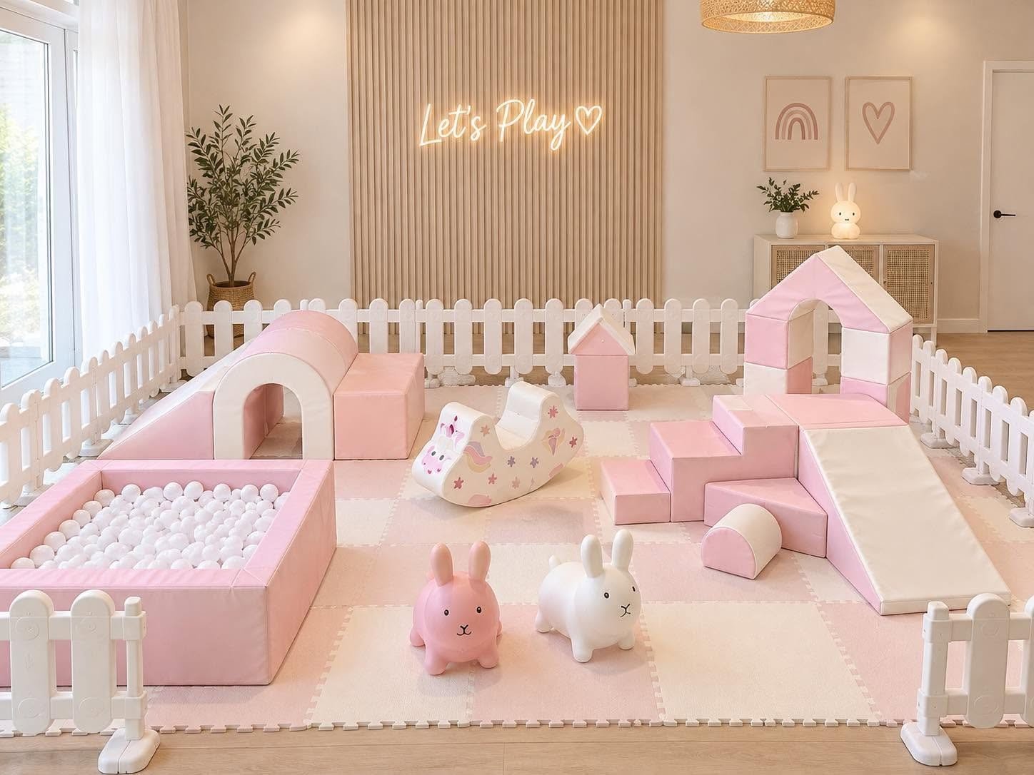 A pastel-colored indoor play area for children with soft pink and white foam structures, a ball pit, bunny and rabbit toys, a slide, and wall decorations featuring a rainbow and a heart, with a sign that reads "Let's Play" in neon lights.