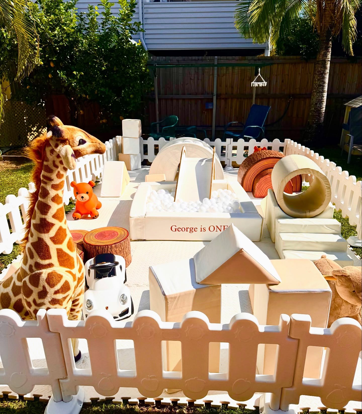 Children's birthday party setup in a backyard with a fenced play area, featuring a plush giraffe toy, a small orange bear toy, foam play shapes, and signs reading 'George is ONE'.