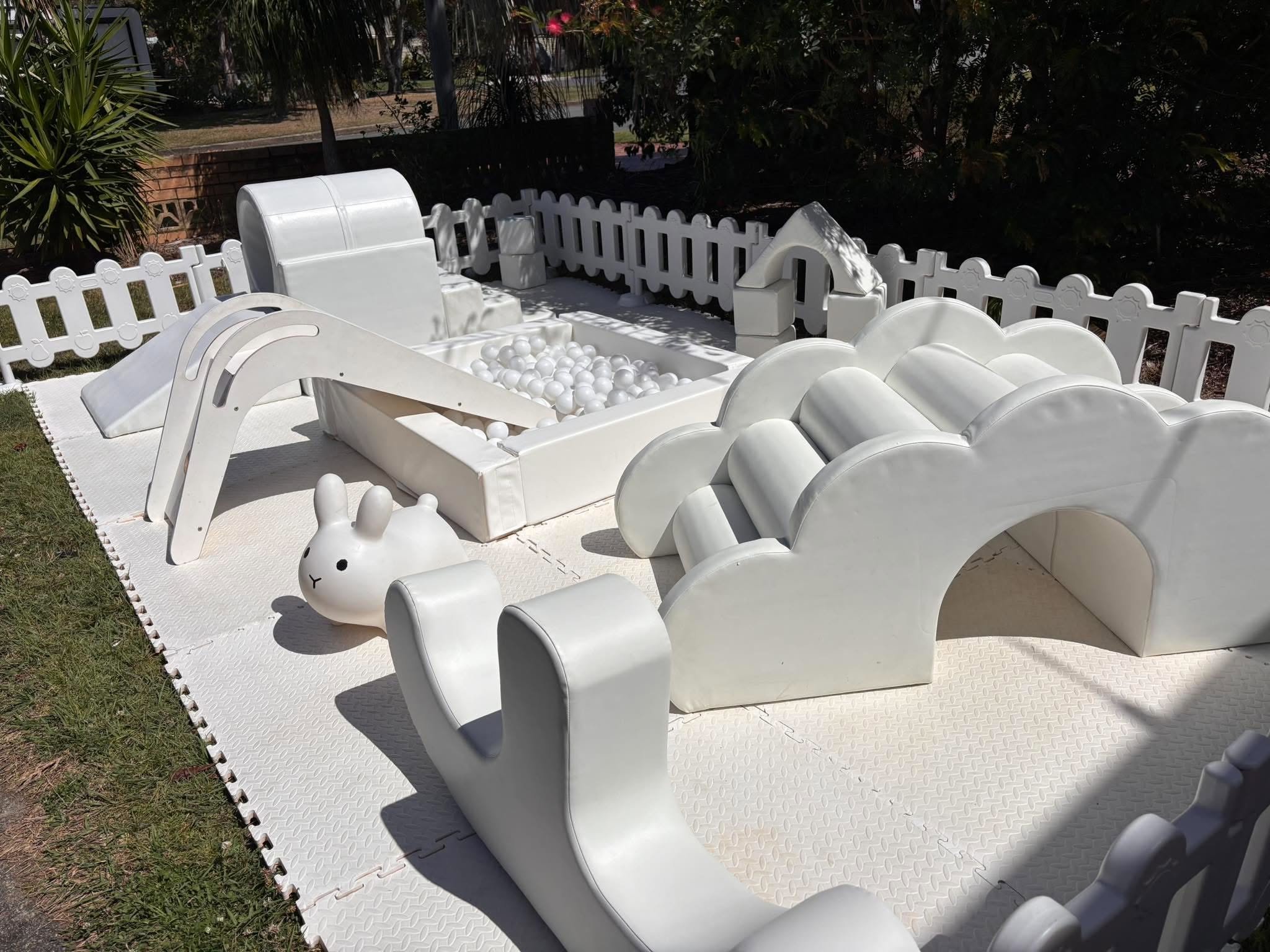 White plastic rabbit-shaped and cloud-shaped children’s playground seats arranged around a rectangular ball pit filled with white balls, set on a white rubberized playground surface, with a white picket fence and trees in the background.