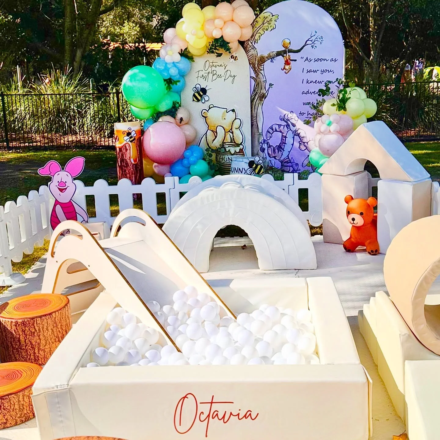 Decorative outdoor setup for a child's first birthday with a Winnie the Pooh theme. Features include balloons, a white picket fence, cartoon cutouts of Winnie the Pooh and Piglet, a ball pit with white balls, a small slide, and a backdrop with illust
