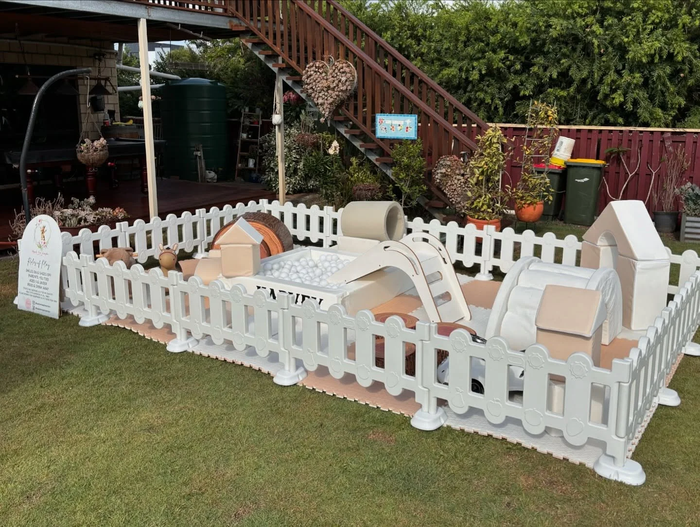 Miniature animal play area surrounded by a white picket fence, with soft flooring, featuring toys and structures for children to play.
