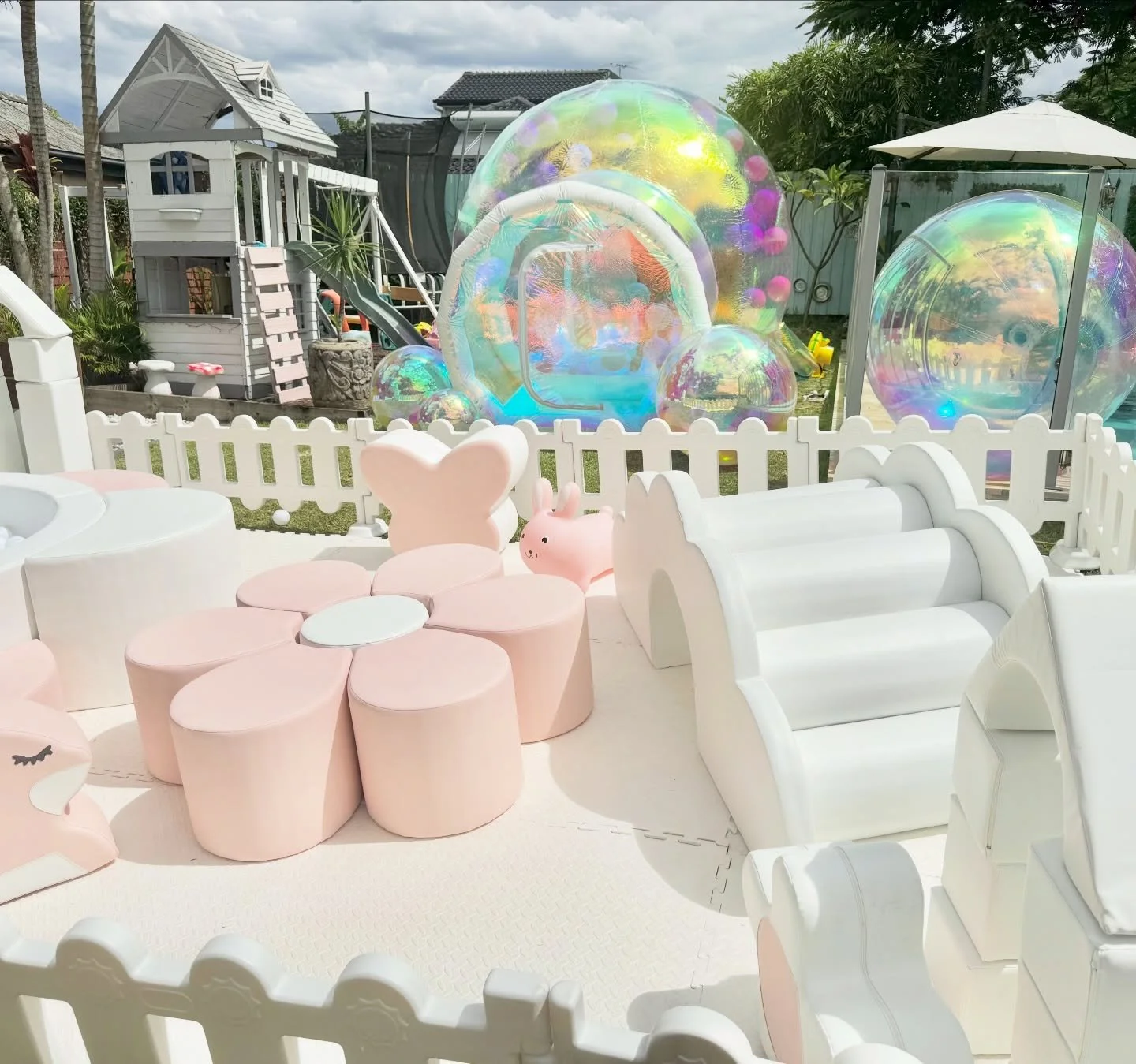 A colorful outdoor play area with pastel pink, white, and peach furniture, including animal-shaped chairs and tables. In the background, there are various bubble-like structures and a small playhouse, along with a playground slide and a trampoline, a