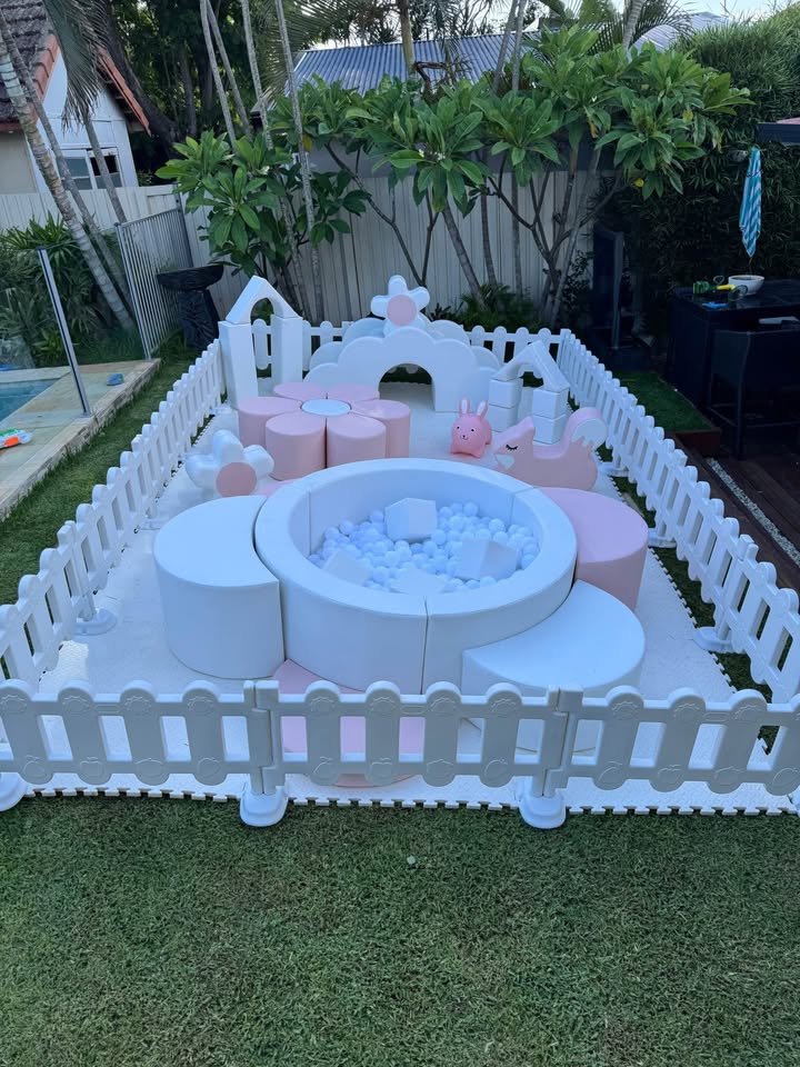 A children's outdoor play area with a white picket fence, featuring a small ball pit with white balls, surrounded by pink and white foam furniture, plush toys, and decorative elements like a bunny and a cloud.