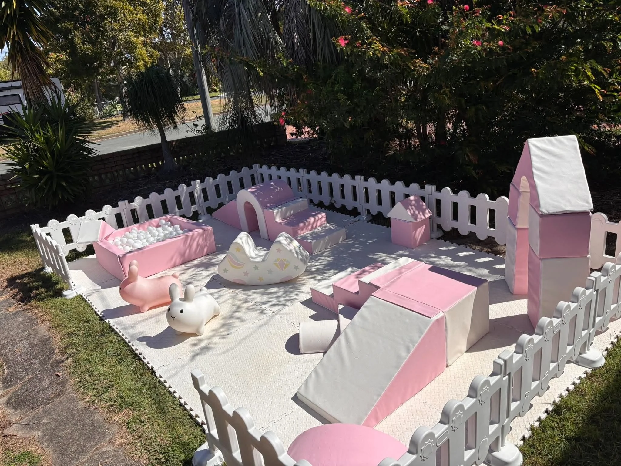 Cute pink and white toy chicken, bunny, and unicorn surrounded by a white picket fence on a patio with outdoor furniture and lush green trees and bushes in the background.