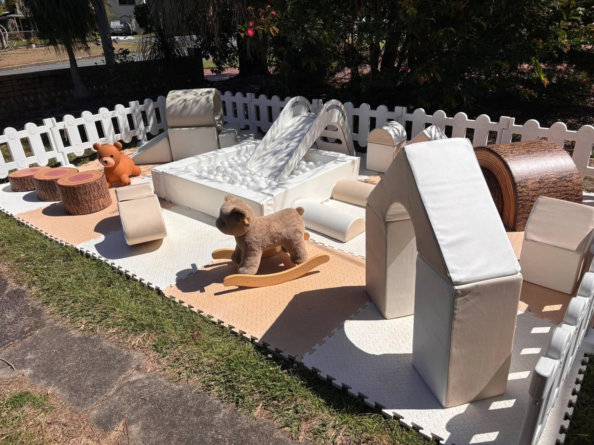 A backyard scene with outdoor play area for children. Features a white plastic slide, a ball pit filled with white balls, a teddy bear, a toy rocking horse, and cut logs. Surrounded by a white picket fence and trees with pink flowers.