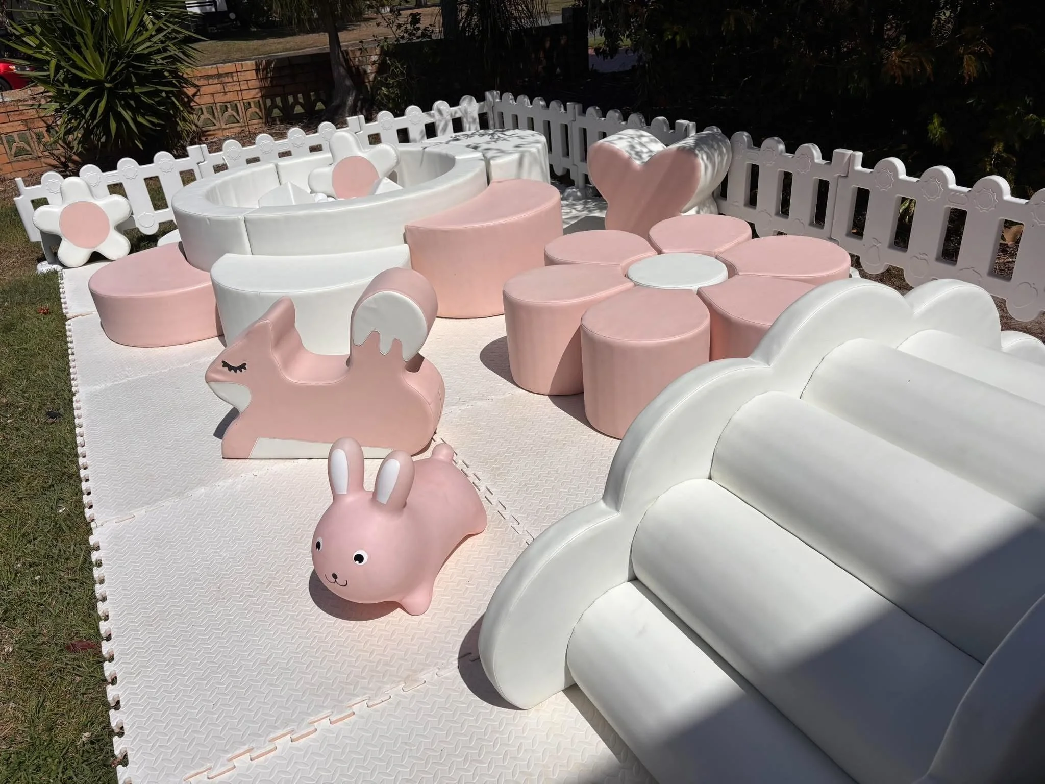 Outdoor children's play area with pink and white bubble-shaped seating, unicorn and heart-shaped furniture, and a white fence, under sunlight.
