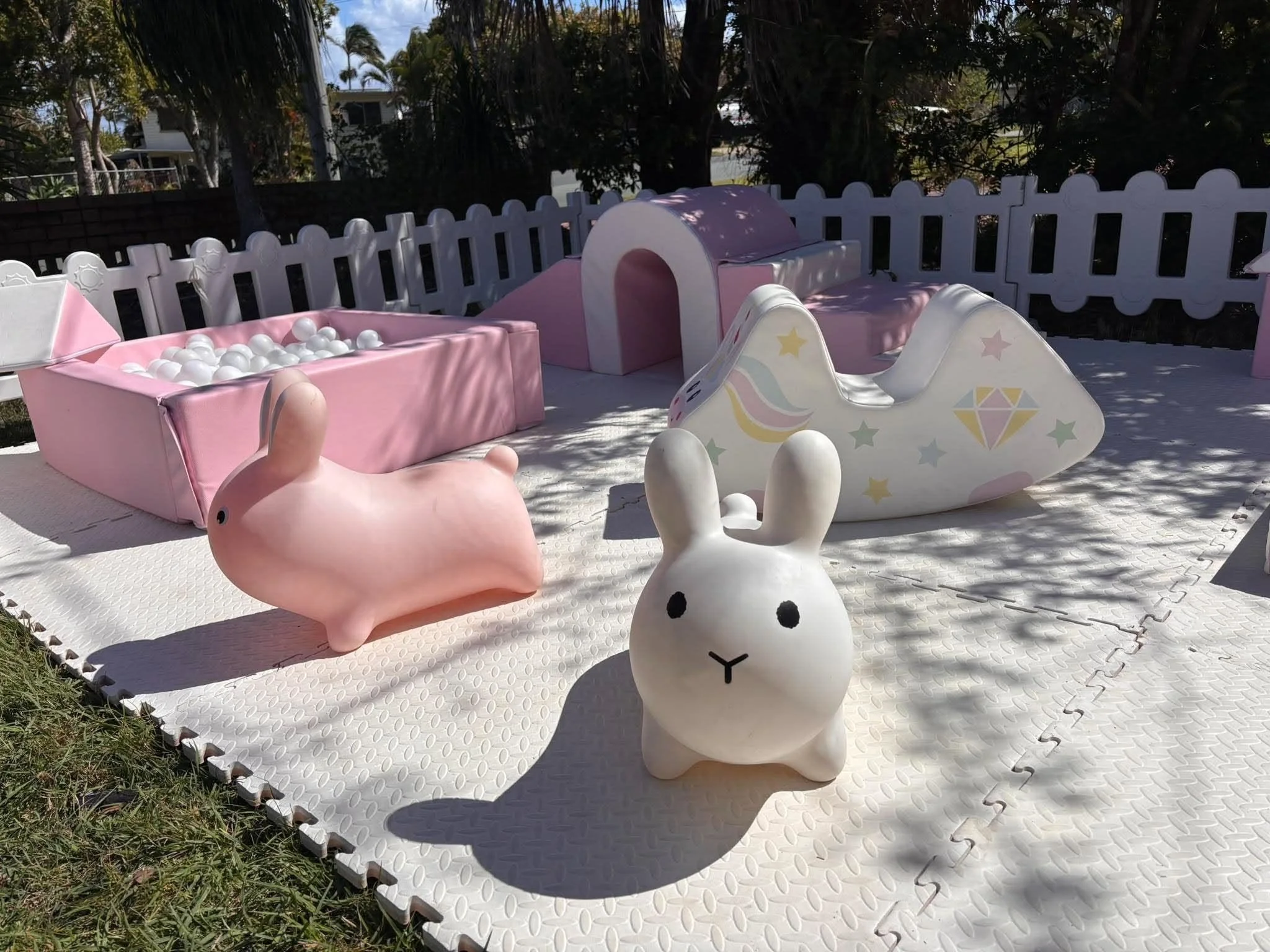 Children's outdoor play area with pastel pink and white unicorn and bunny-shaped toys, surrounded by a white picket fence and greenery in the background.