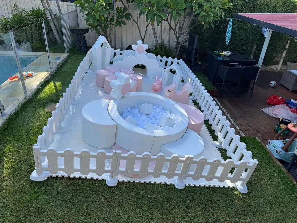 A small, enclosed outdoor play area with a white picket fence. Inside is a pink and white pastel-colored playhouse with a ball pit filled with white balls. There are plush toys around the playhouse, including a pig and a unicorn. The area is on a gra
