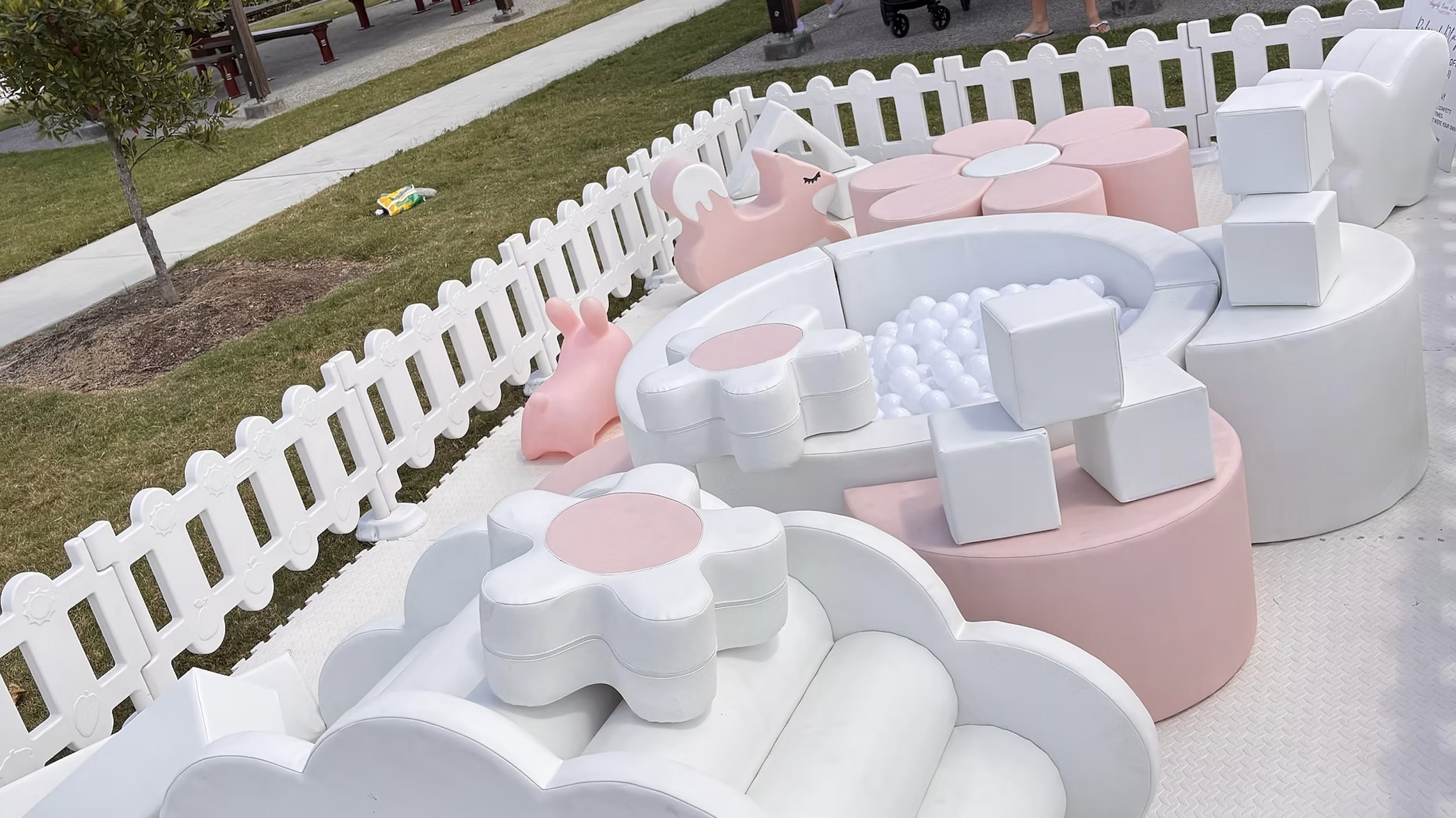 Pink and white outdoor furniture with a ball pit, shaped like flowers with bunny and swan decorations, surrounded by a white picket fence, situated on a grassy area with a sidewalk nearby.