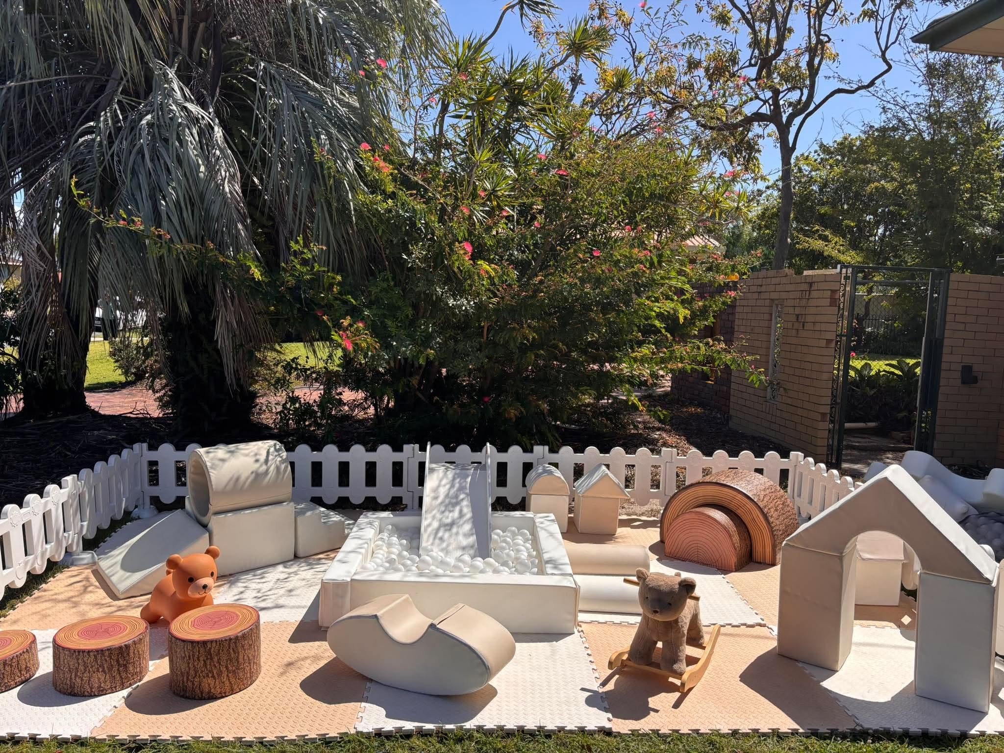 Outdoor play area with white plastic furniture, teddy bear and toy animal, surrounded by trees and a brick wall