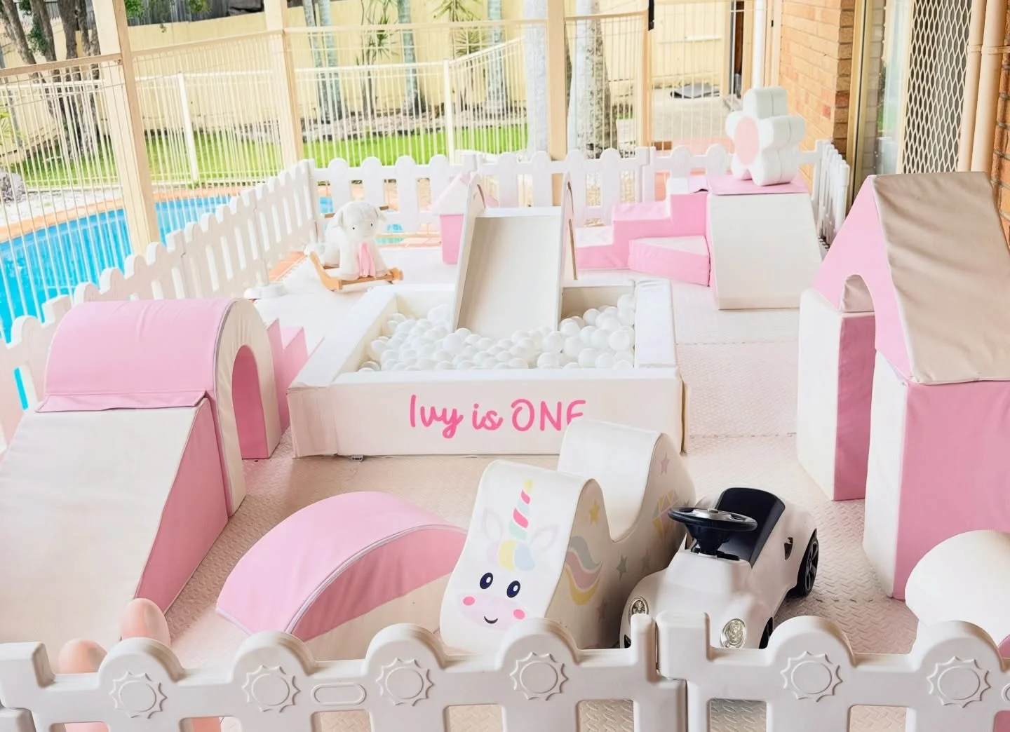 Colorful indoor play area for children with pink and white soft structures, ball pit, toy car, and unicorn-themed decorations.