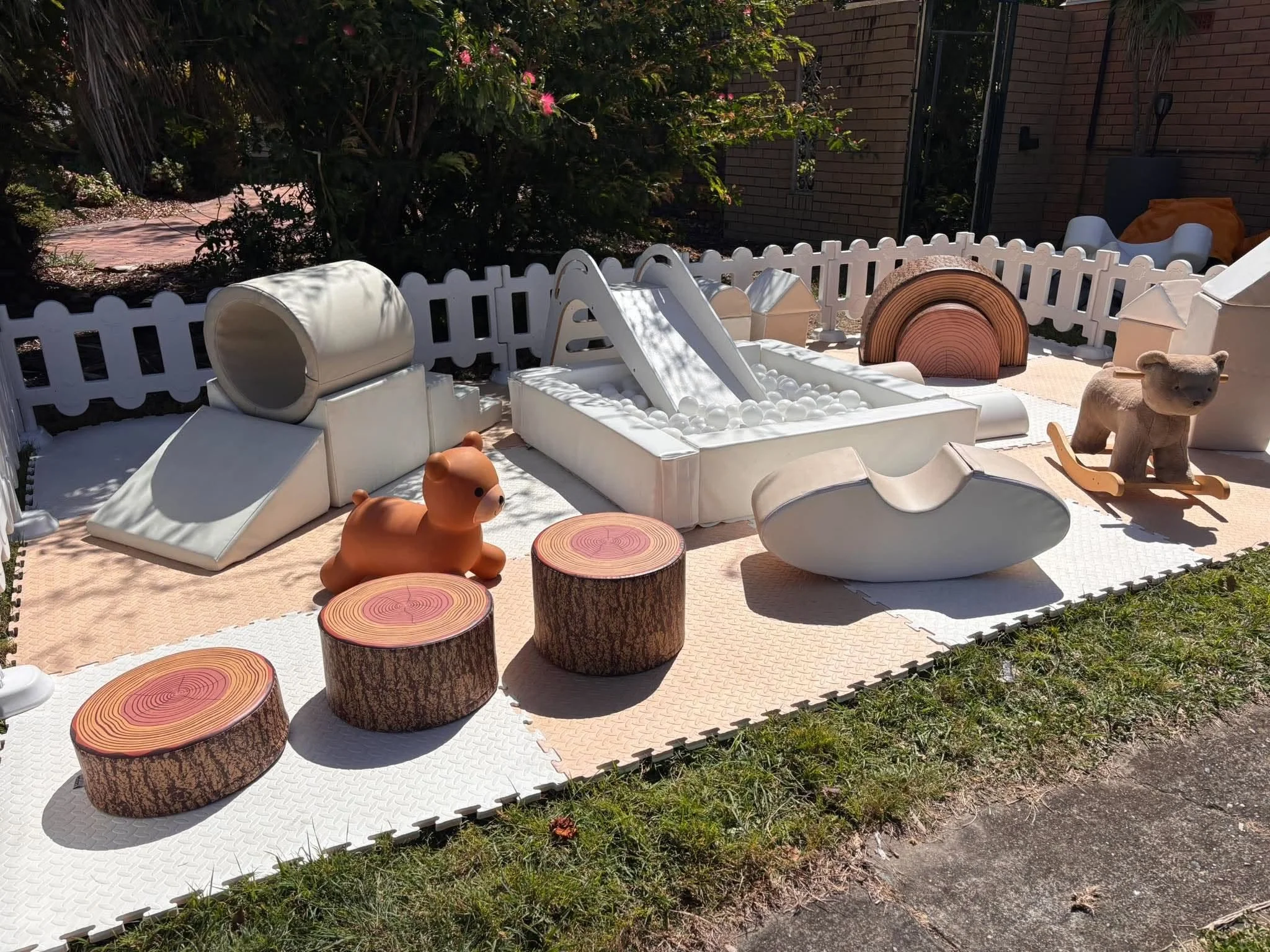 A small outdoor play area with white mini playground equipment including a slide, tunnel, and ball pit, surrounded by a white picket fence and decorated with tree stump seating and a plush bear toy, set against a brick house and lush greenery in the 