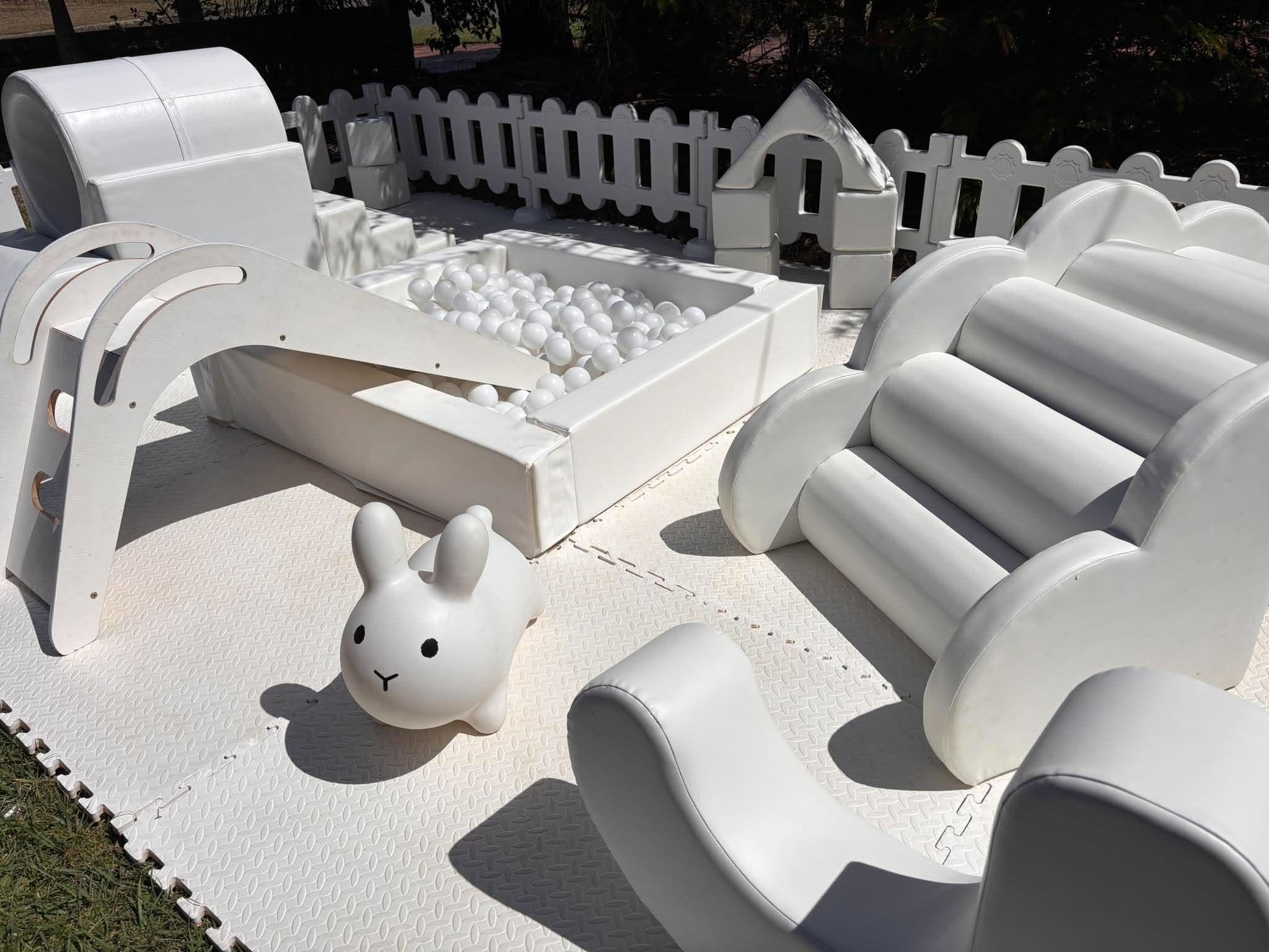Outdoor children's play area with white seating, a bunny-shaped decoration, and a ball pit filled with white balls, enclosed by a white picket fence.