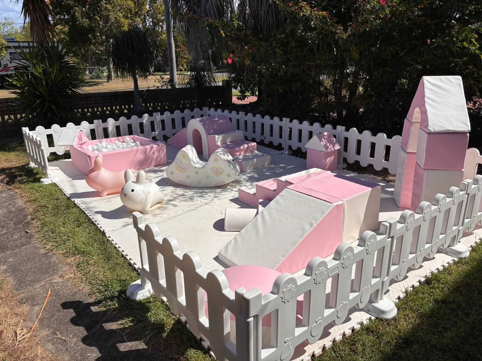 Outdoor play area with pink and white toy furniture, a ball pit, rabbit-shaped decoration, surrounded by a white picket fence, trees, and a grassy area.