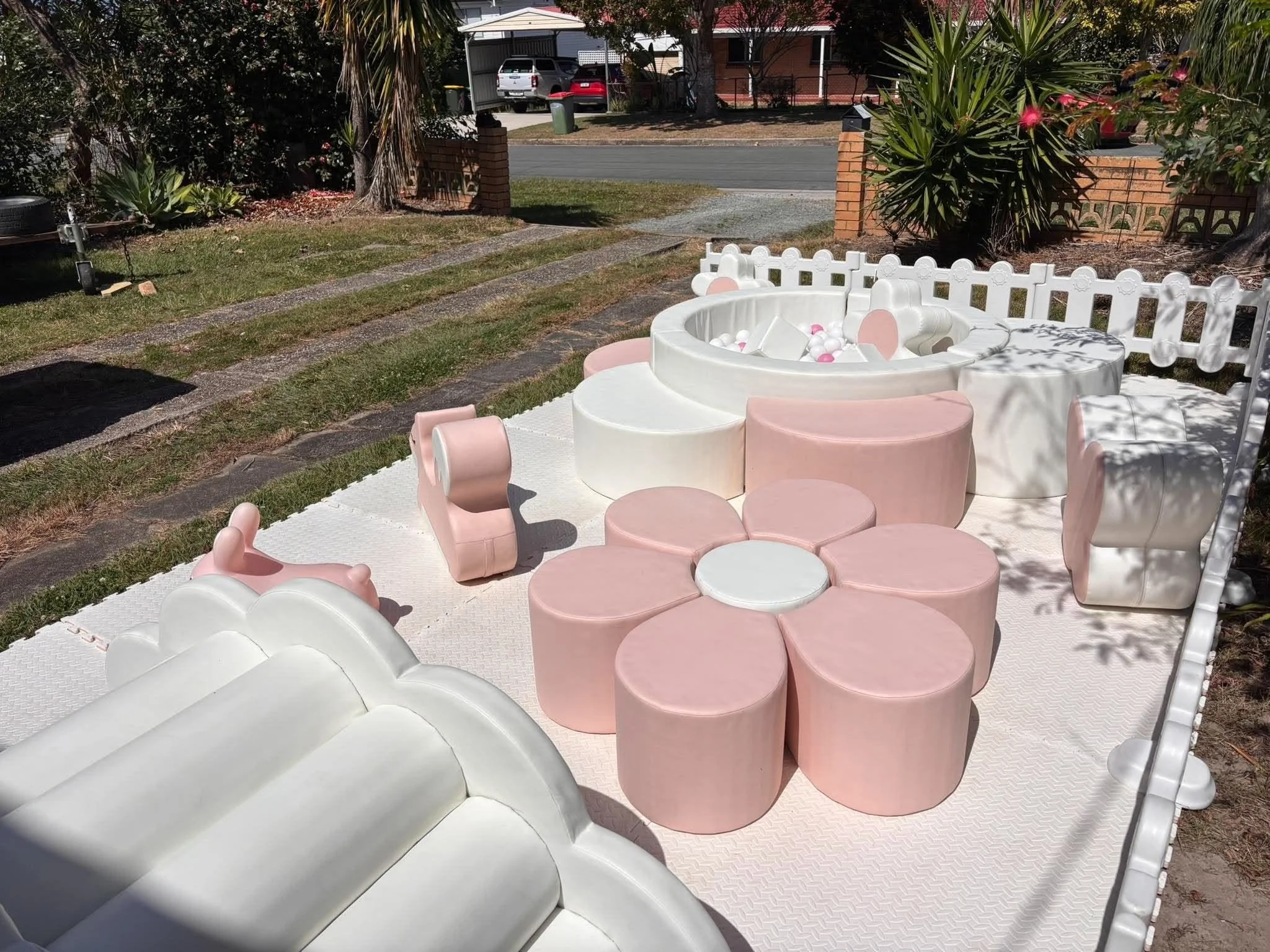 A backyard scene with a small plastic pool filled with white and pink balls, surrounded by pink and white chairs. There are trees and bushes in the background, and a driveway with parked cars in front of a house.