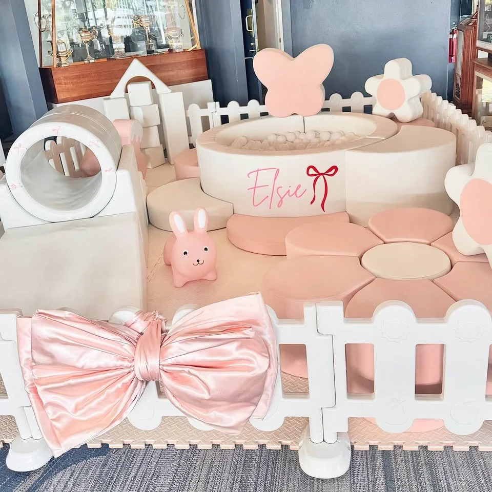 Decorated play area with pink and white theme, featuring a small pool with a butterfly shape, plush bunny toy, large pink satin bow, and various animal-shaped pillows and decorations. The name 'Elsie' is written on the side of the pool.
