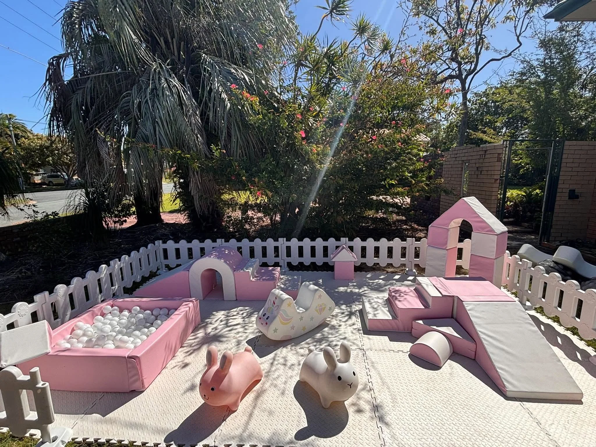 A backyard play area with pink cushioned benches, white plastic bunny and unicorn figures, a small ball pit filled with white balls, and various toys. Tall trees and a brick wall surround the area, with clear blue sky overhead.