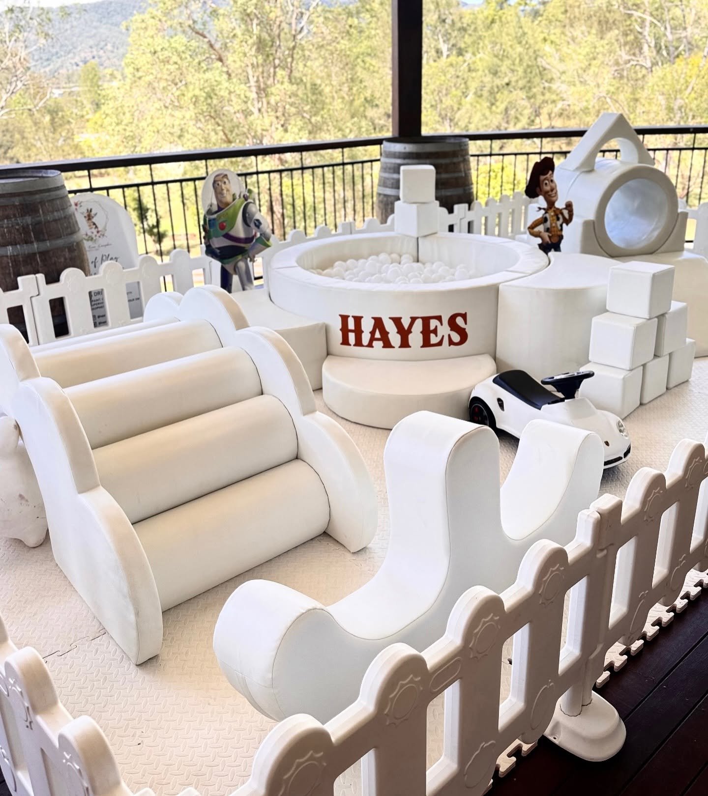 A Toy Story-themed play area featuring white toy furniture, a small toy car, two figurines of Buzz Lightyear and Woody, a ball pit with the name 'HAYES' on it, and a white plastic fence surrounding the area, with trees and a mountain visible outside in the background.