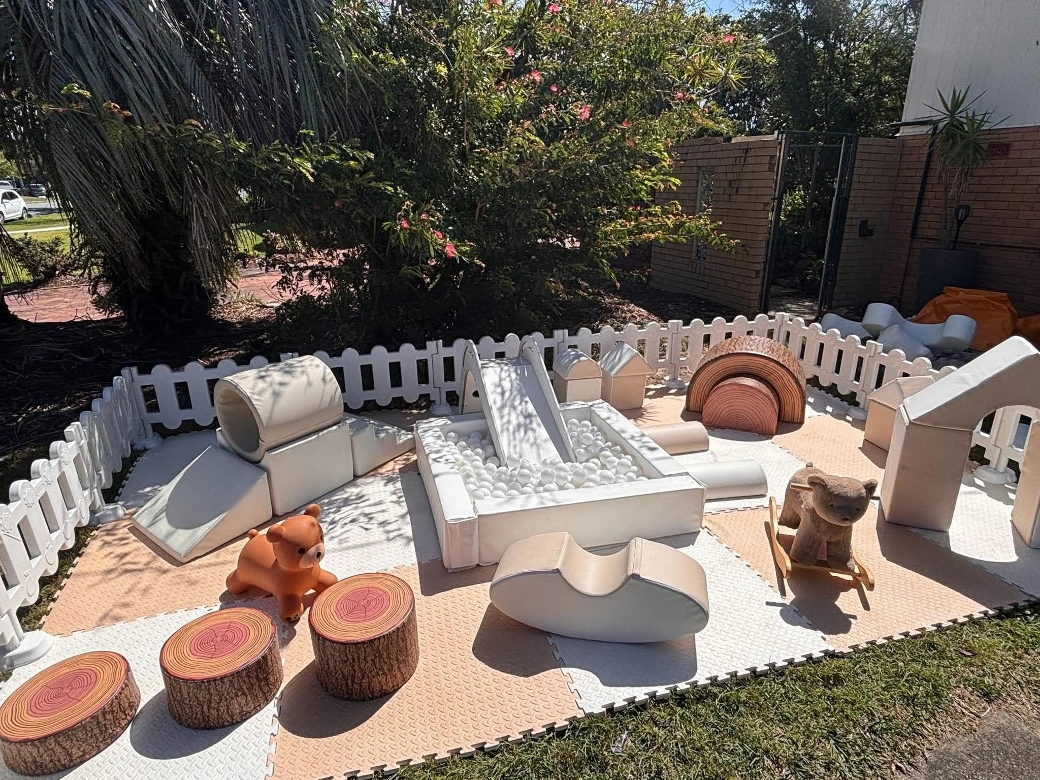 Outdoor patio area with white furniture, animal-shaped decor, and tree stumps used as tables, surrounded by a white fence and greenery.