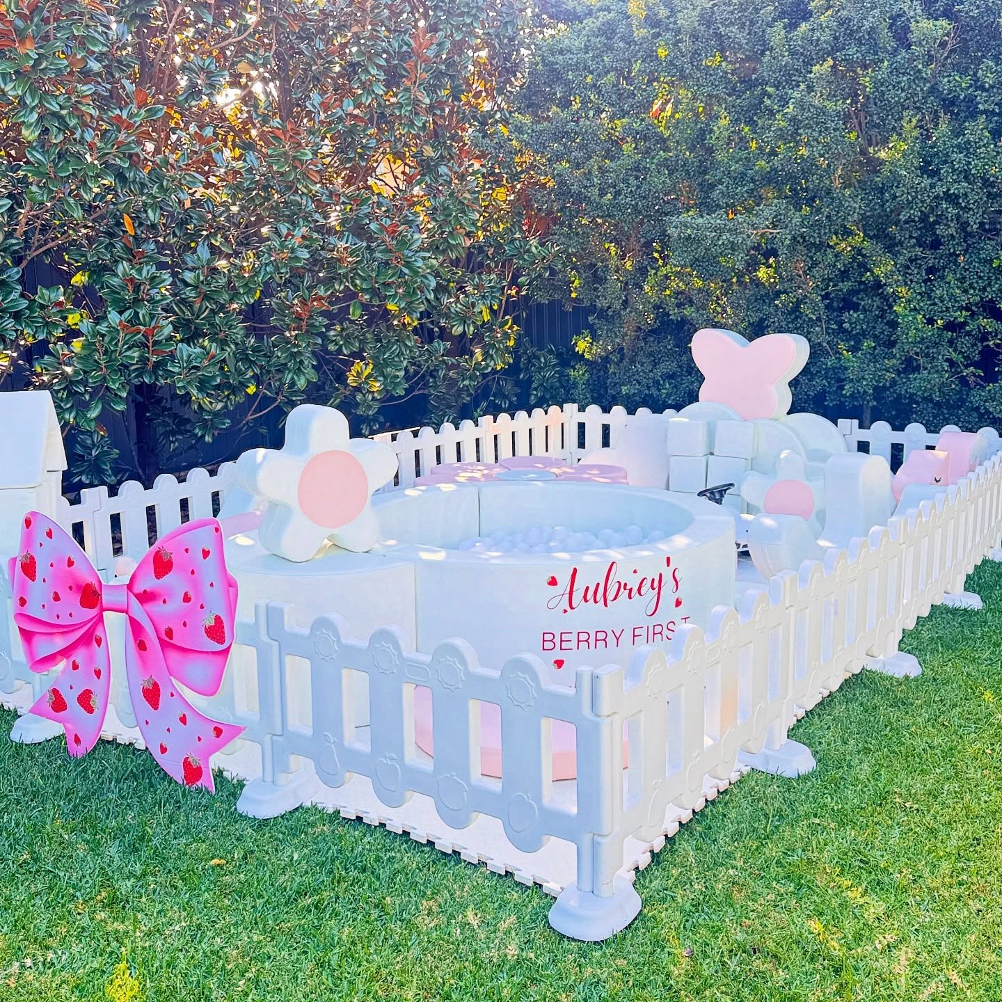 Decorative outdoor play area for a child's first birthday party, enclosed by a white picket fence. The area has pink, white, and pastel-colored decorative elements like large flowers and a bow, with artificial grass underneath. There is a sign that r