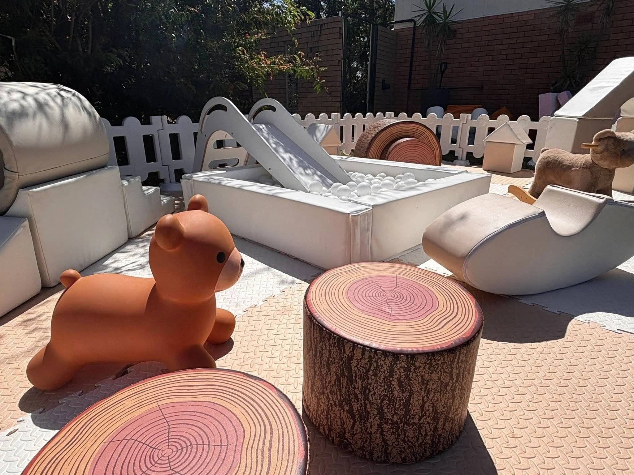 Outdoor patio area with white fence, wooden log tables, rubber duck, and various toys, shaded by trees.