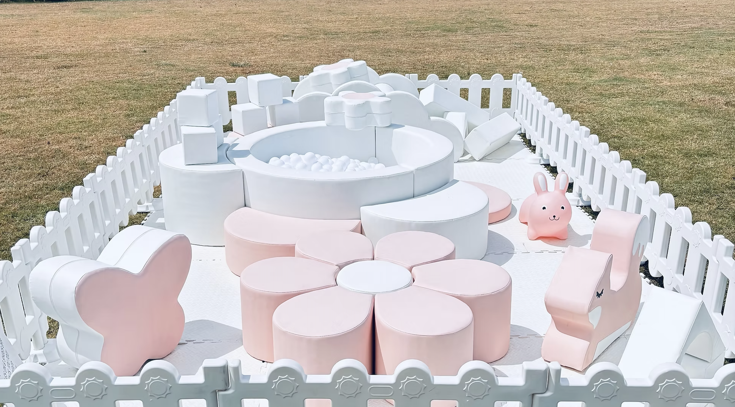 A pink and white children's play area outdoors with a bunny-shaped seat, a mini pool, and various soft blocks enclosed by a white picket fence.