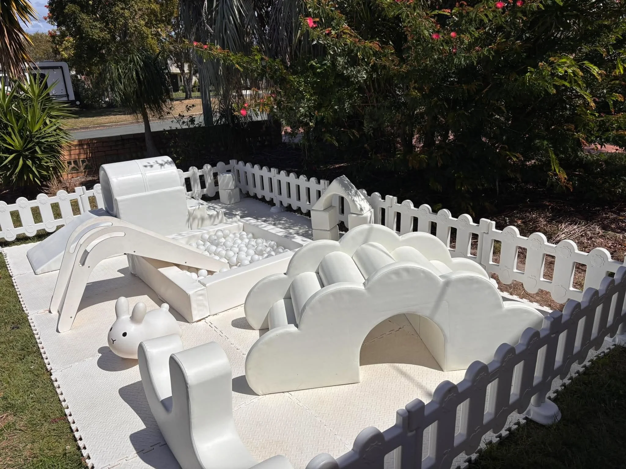 Outdoor scene featuring white plastic toys, including a bunny-shaped container and balls, surrounded by a small white picket fence on a large white mat, with lush green bushes and trees in the background, and a clear sunny sky.