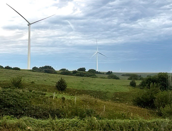 Rolling green hills in Nebraska with several wind turbines under a cloudy sky.