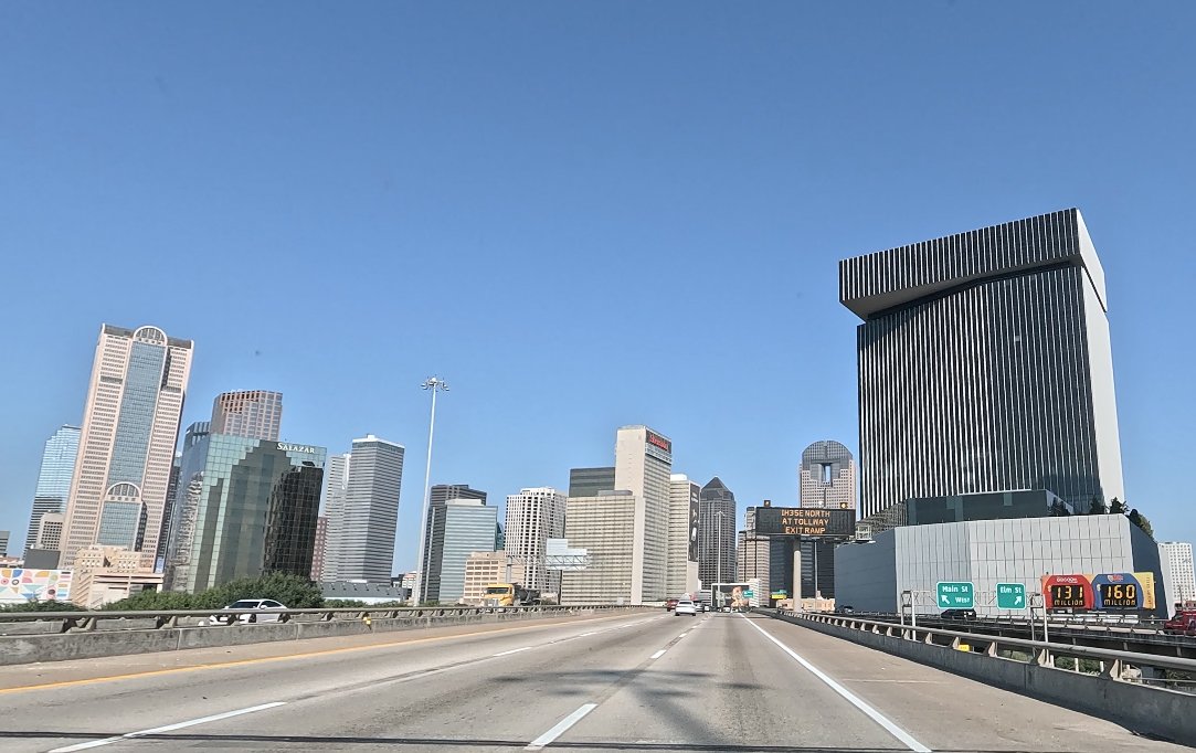 View of the Dallas skyline from an elevated highway, with downtown buildings beneath a clear blue sky and traffic signs along the roadway.