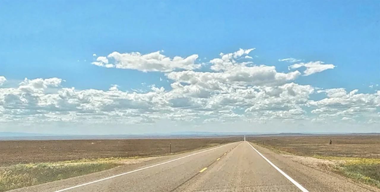A long, empty highway stretches toward the horizon through a vast, open landscape beneath a bright blue sky filled with scattered clouds.