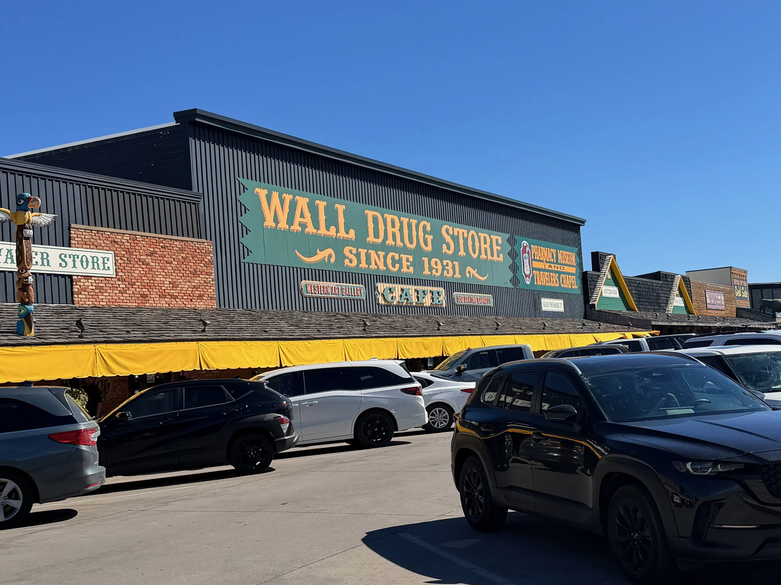 Green and gold Wall Drug Store sign, "Since 1931," above a parking lot full of cars.