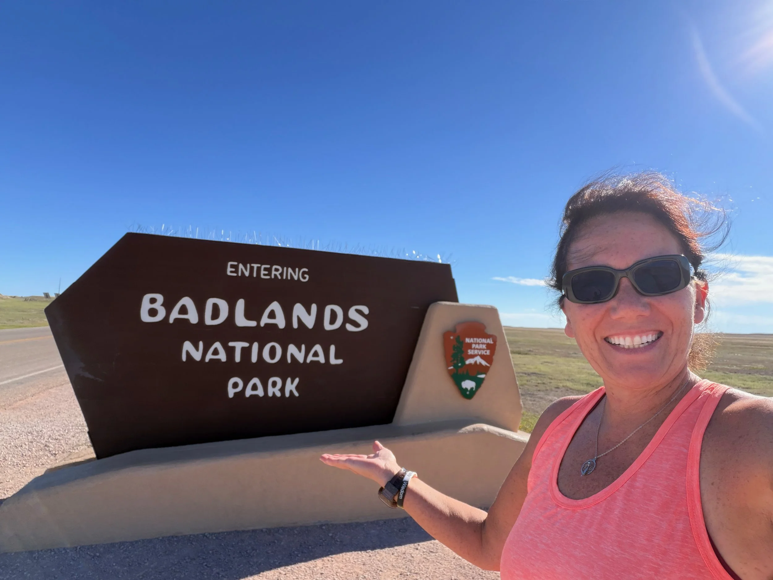 Smiling woman in sunglasses gesturing toward the "Entering Badlands National Park" sign.