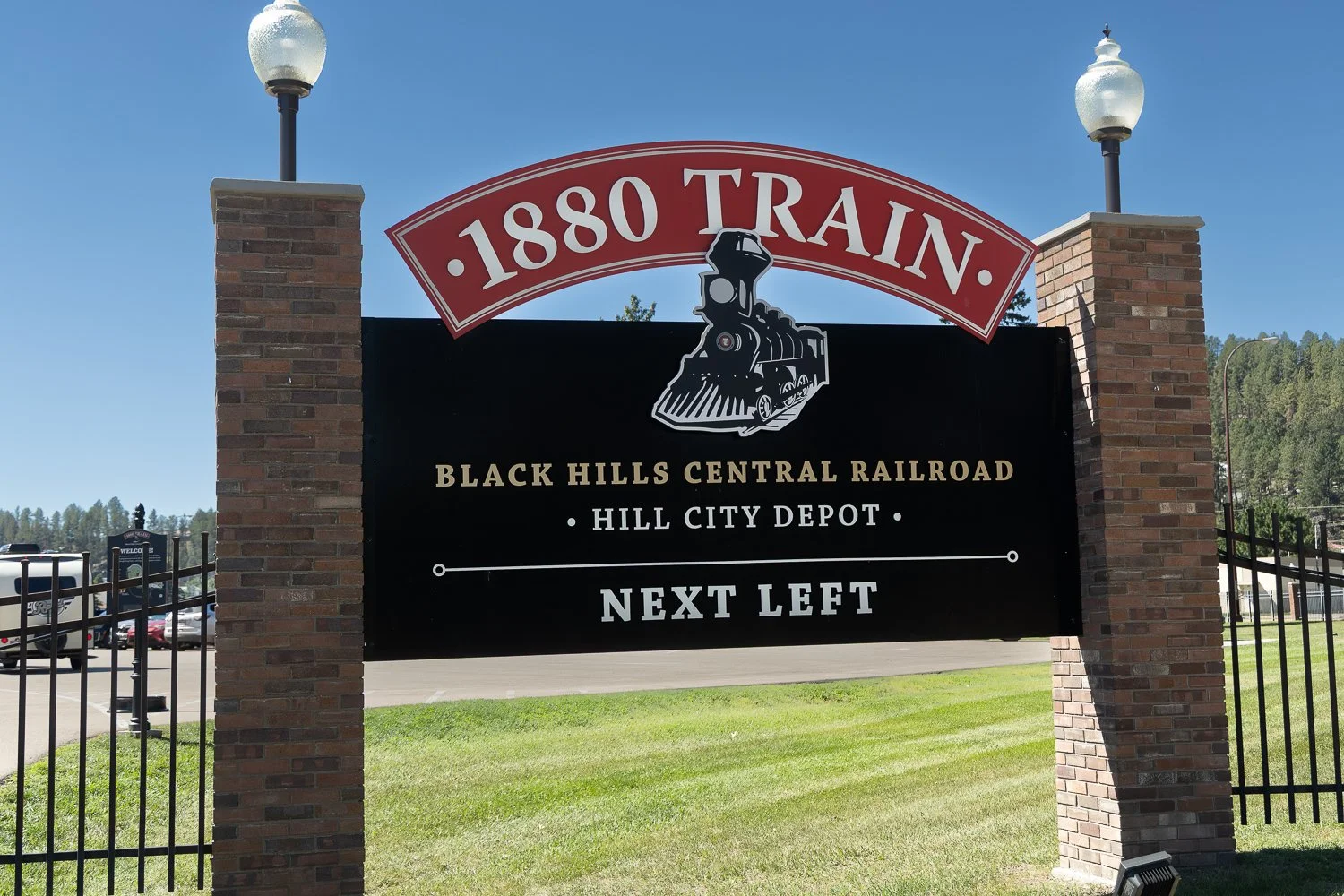 Brick-pillared "1880 Train, Black Hills Central Railroad, Hill City Depot" sign under a clear blue sky.