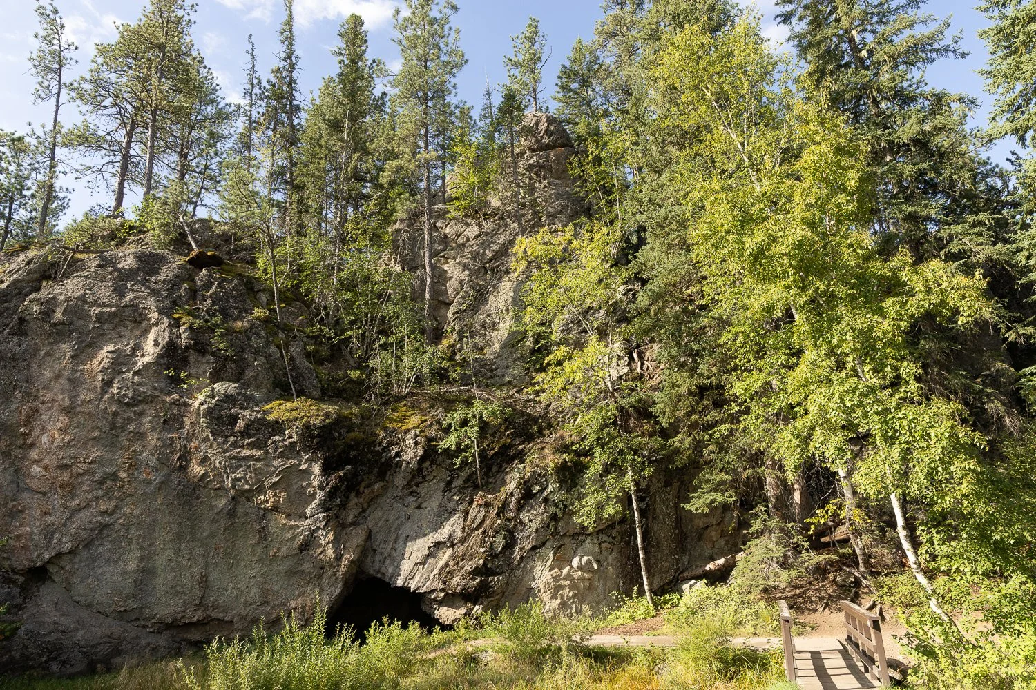 Rocky outcrop surrounded by pine and aspen trees, with a wooden footbridge leading toward a cave opening.