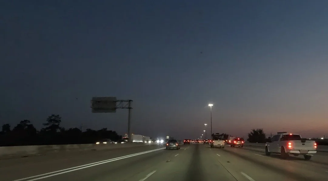 Highway traffic at dawn, with vehicle lights glowing beneath a dark blue sky and a faint glow of morning light on the horizon.