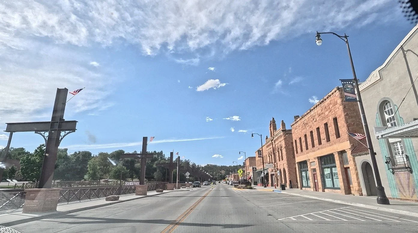 A sunny downtown street lined with historic sandstone buildings, American flags, decorative streetlights, and a pedestrian walkway beside a tree-lined creek.