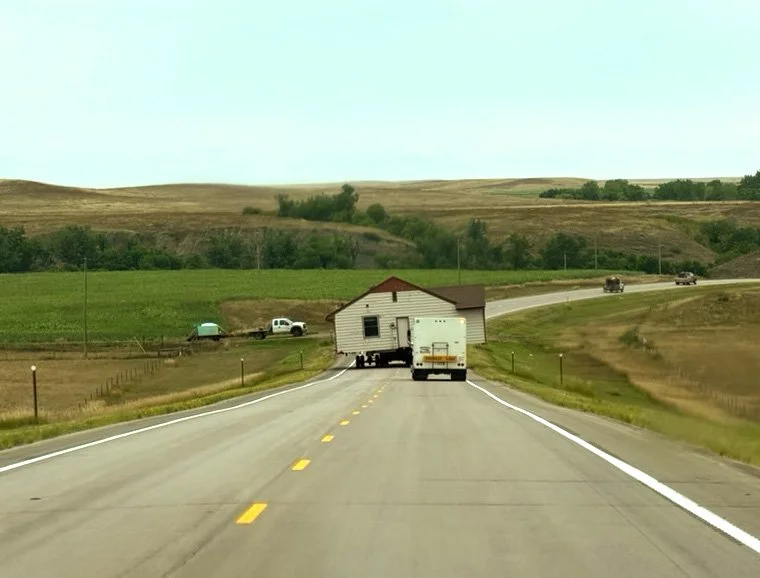 Highway winding through rolling Nebraska farmland with a farm building and an oversized load truck ahead.