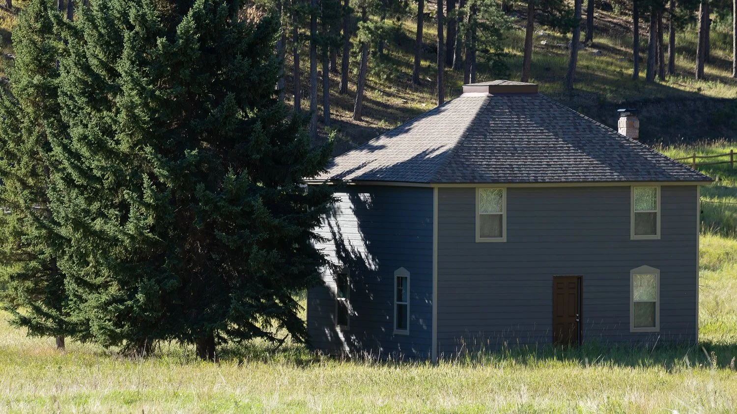 Gray two-story house beside tall evergreens in a grassy meadow at Addie Camp, seen from the 1880 Train.