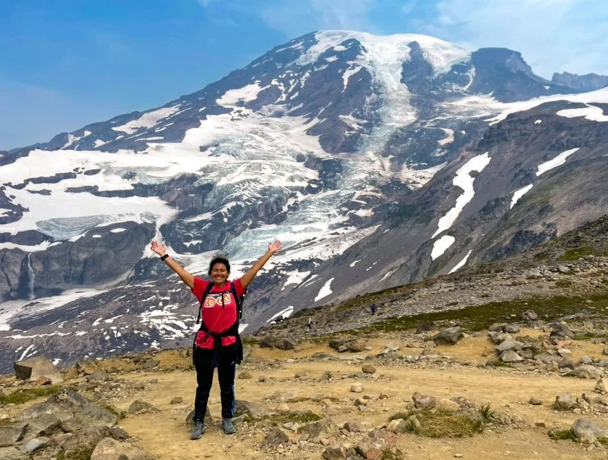 Julie stands with her arms raised joyfully while hiking on Mount Rainier, with a vast, snow-covered mountain landscape behind her.