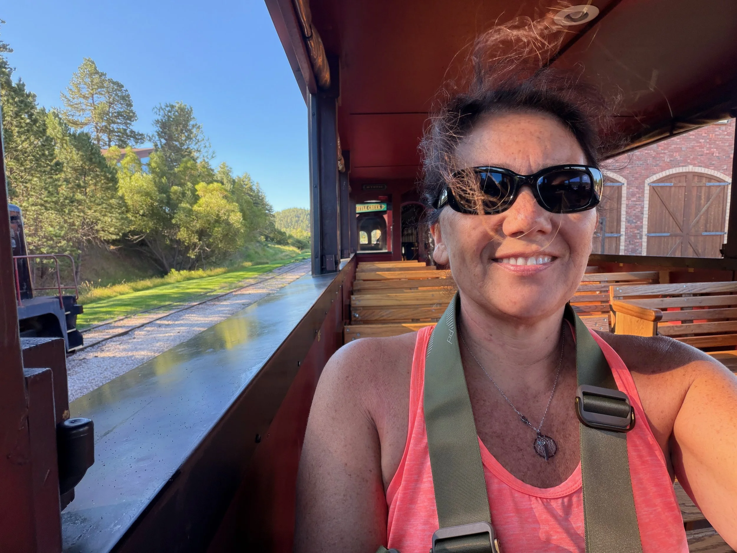 Woman in sunglasses smiling in the 1880 Train's open-air observation car, with wooden benches and the tracks behind her.