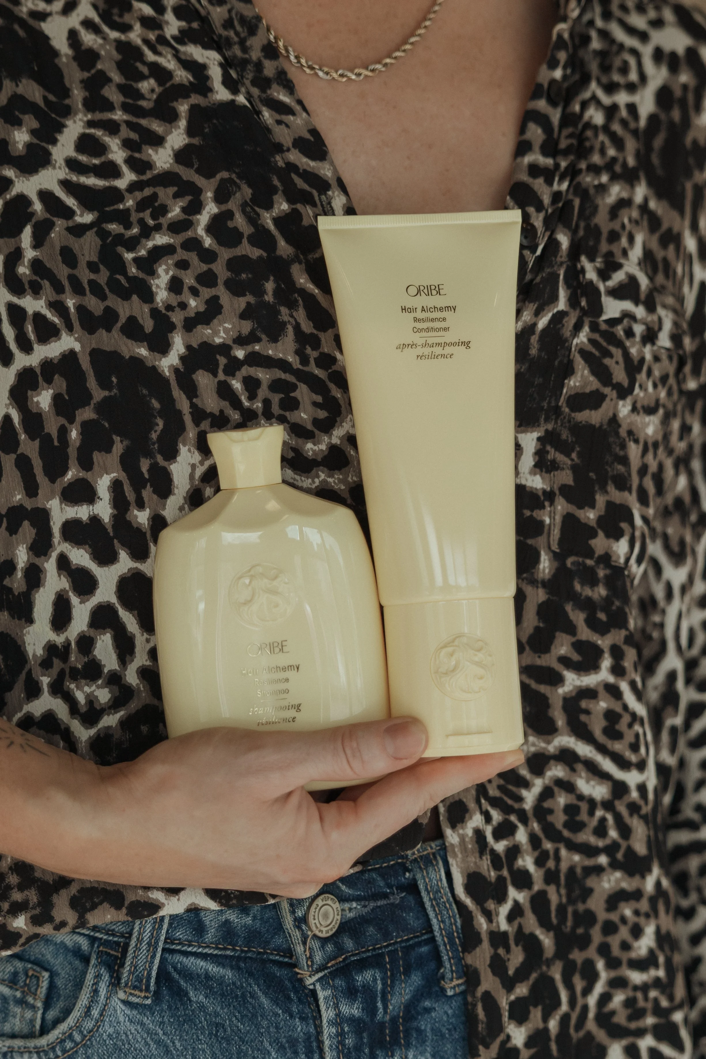 A person wearing a leopard print shirt and jeans holding two bottles of Oribe Hair Alchemy Resilience Conditioner and Shampoo in front of their chest.