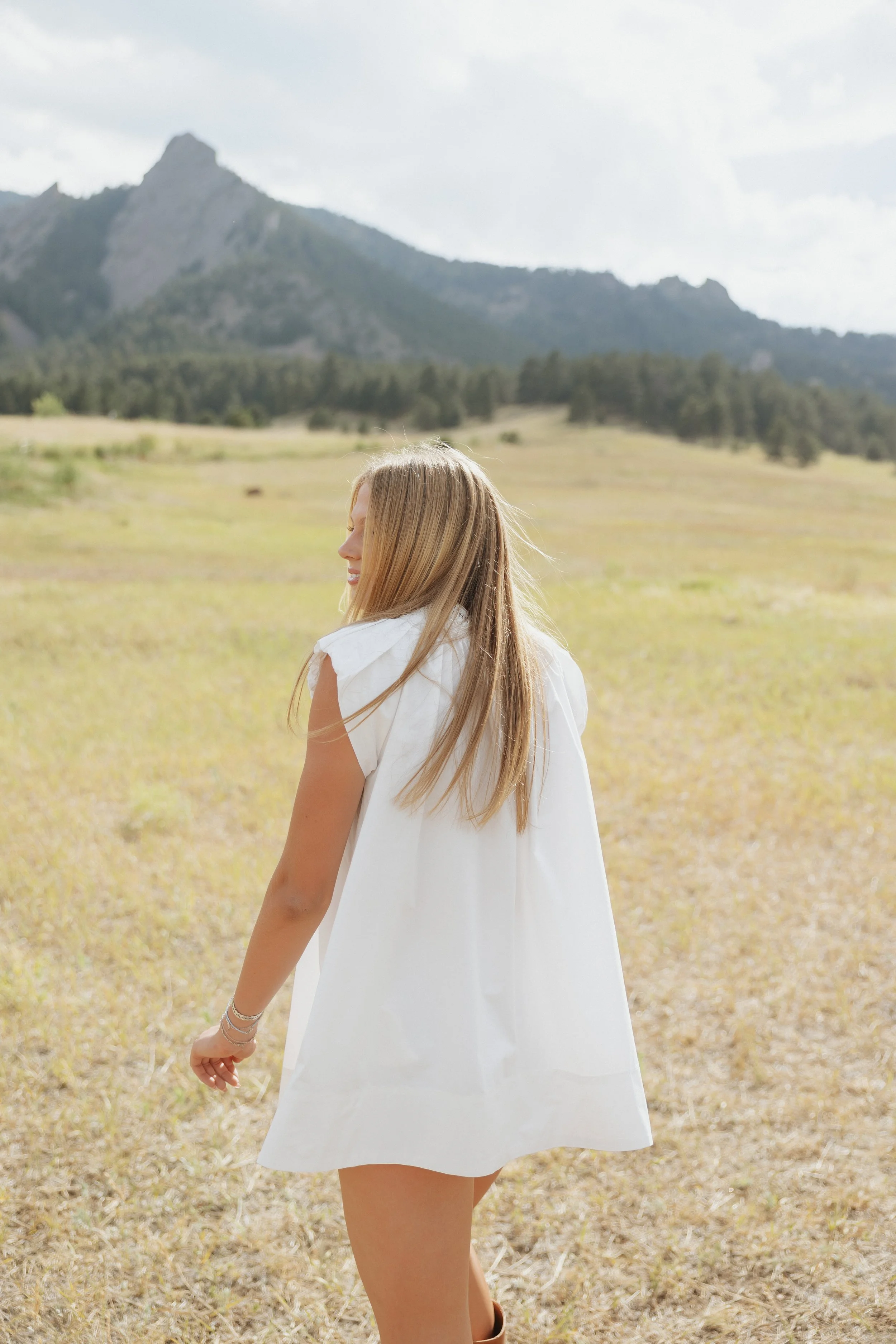 A young woman with long blonde hair in a white dress standing in an open field with mountains in the background, looking to her left with a slight smile.